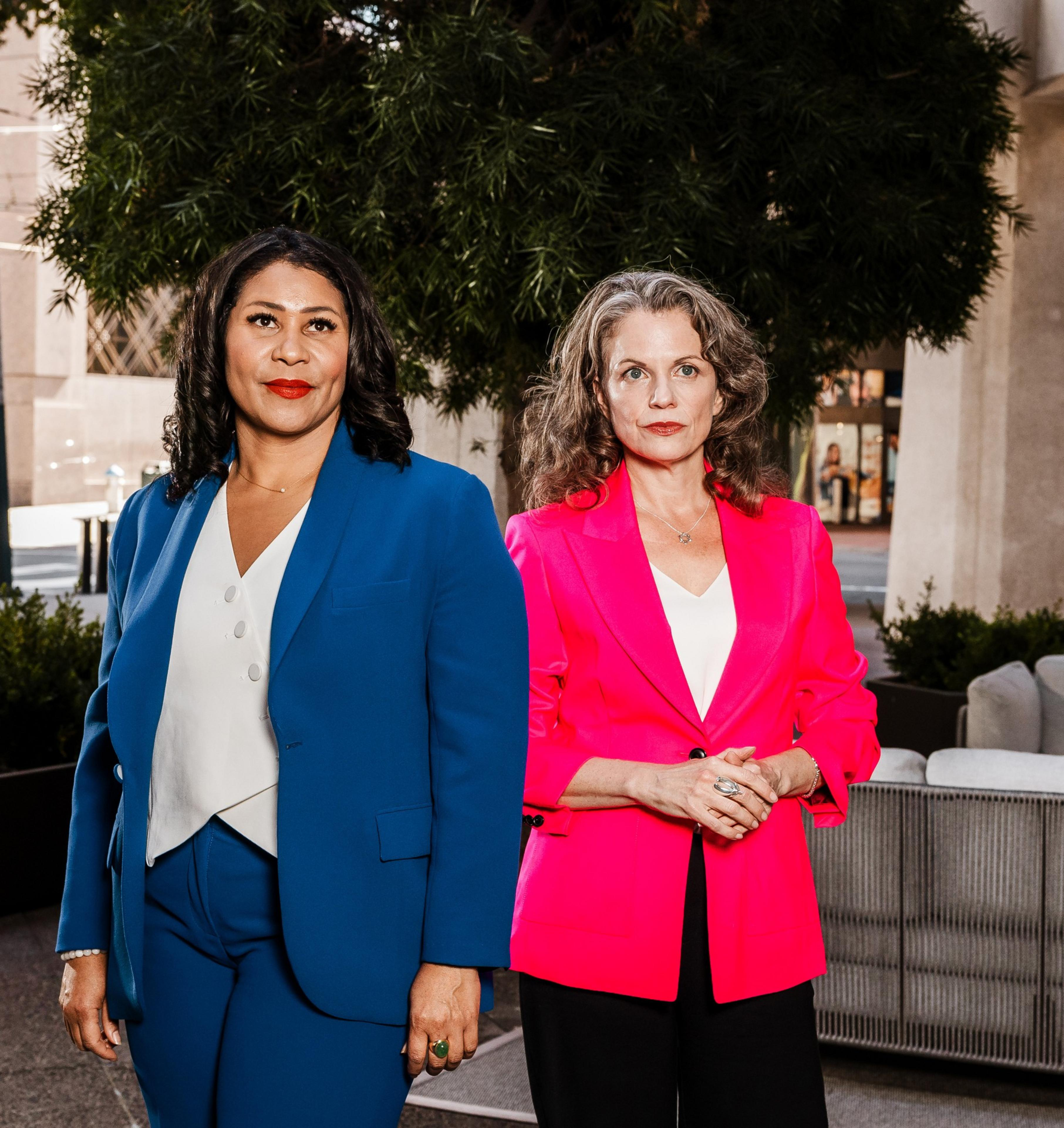 Two women stand side by side outdoors; the one on the left wears a blue suit and white blouse, while the one on the right wears a bright pink blazer over a white top.