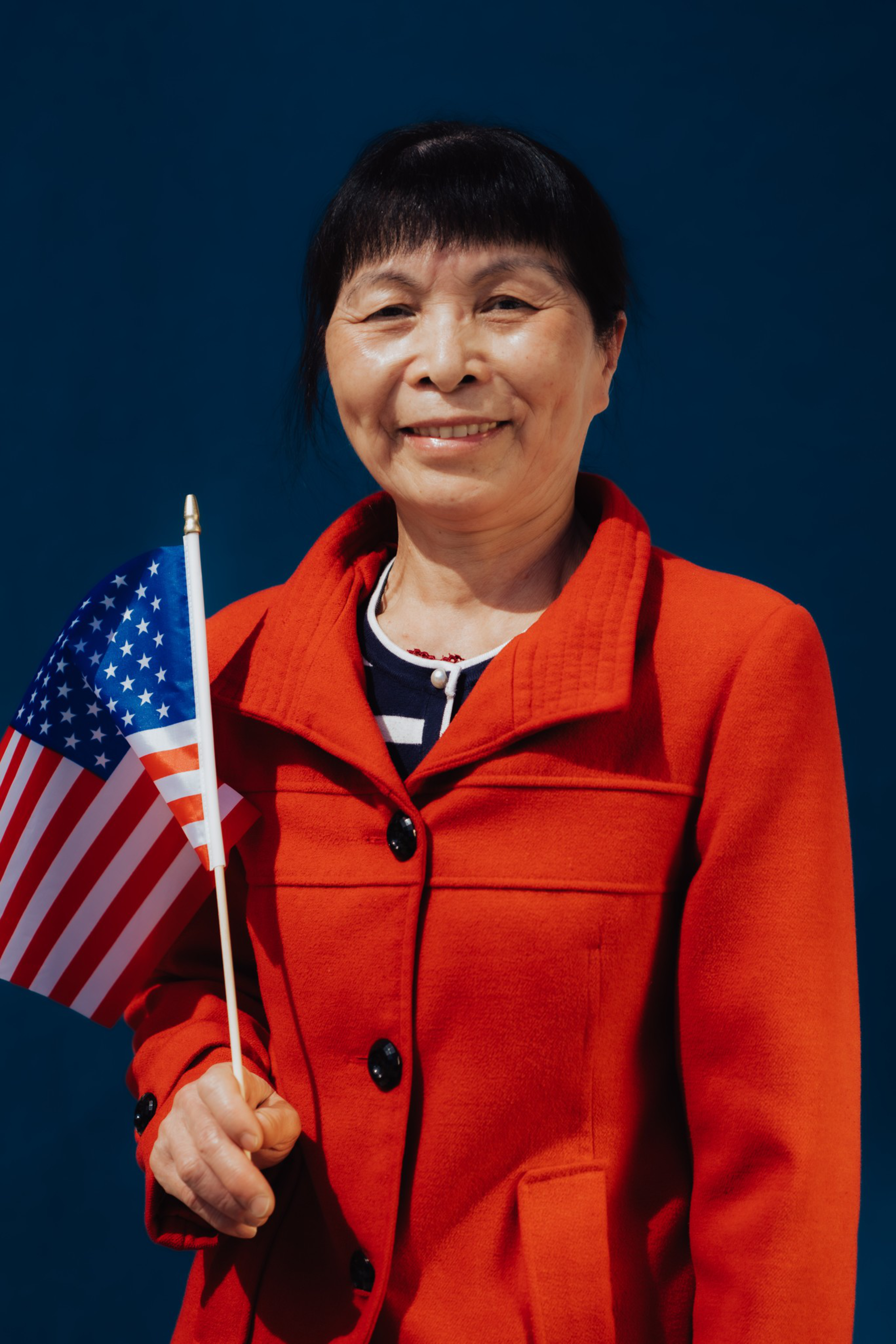 A smiling woman in a red coat holds a small American flag against a solid dark blue background.