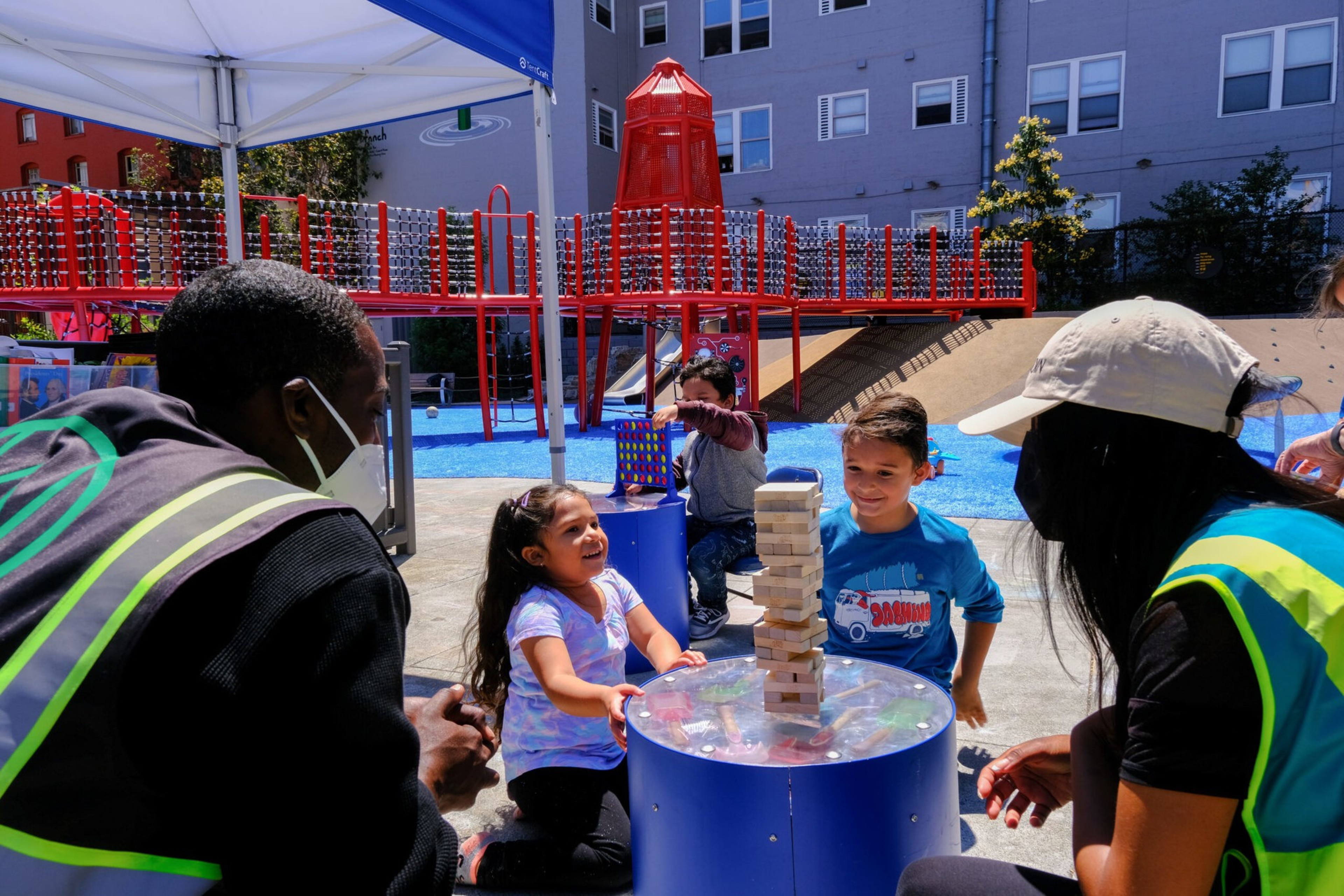Two children and two adults play a large Jenga game outdoors near a red playground structure on a sunny day.