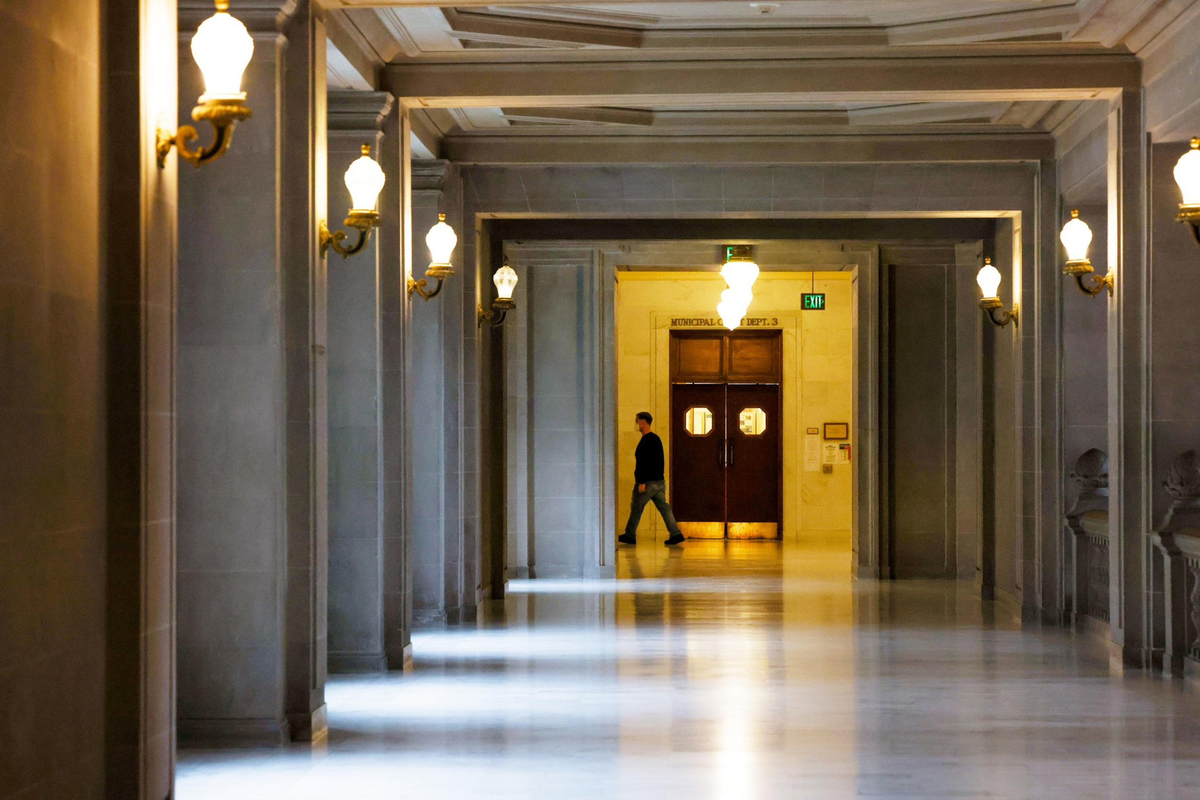 A person walks down a long, marble-floored corridor lined with glowing wall lamps, heading toward wooden double doors under a large ceiling light.