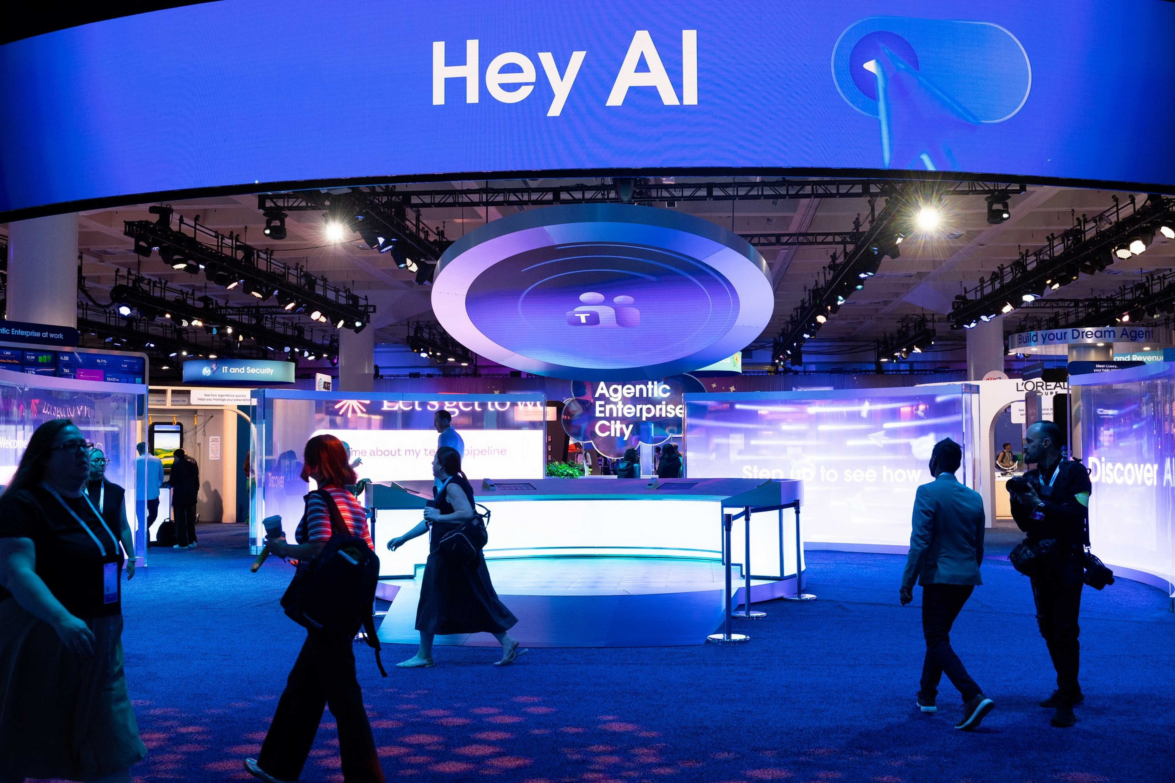 A tech expo booth features a large “Hey AI” sign and “Agentic Enterprise City” display, with people walking and interacting around it under bright lights.