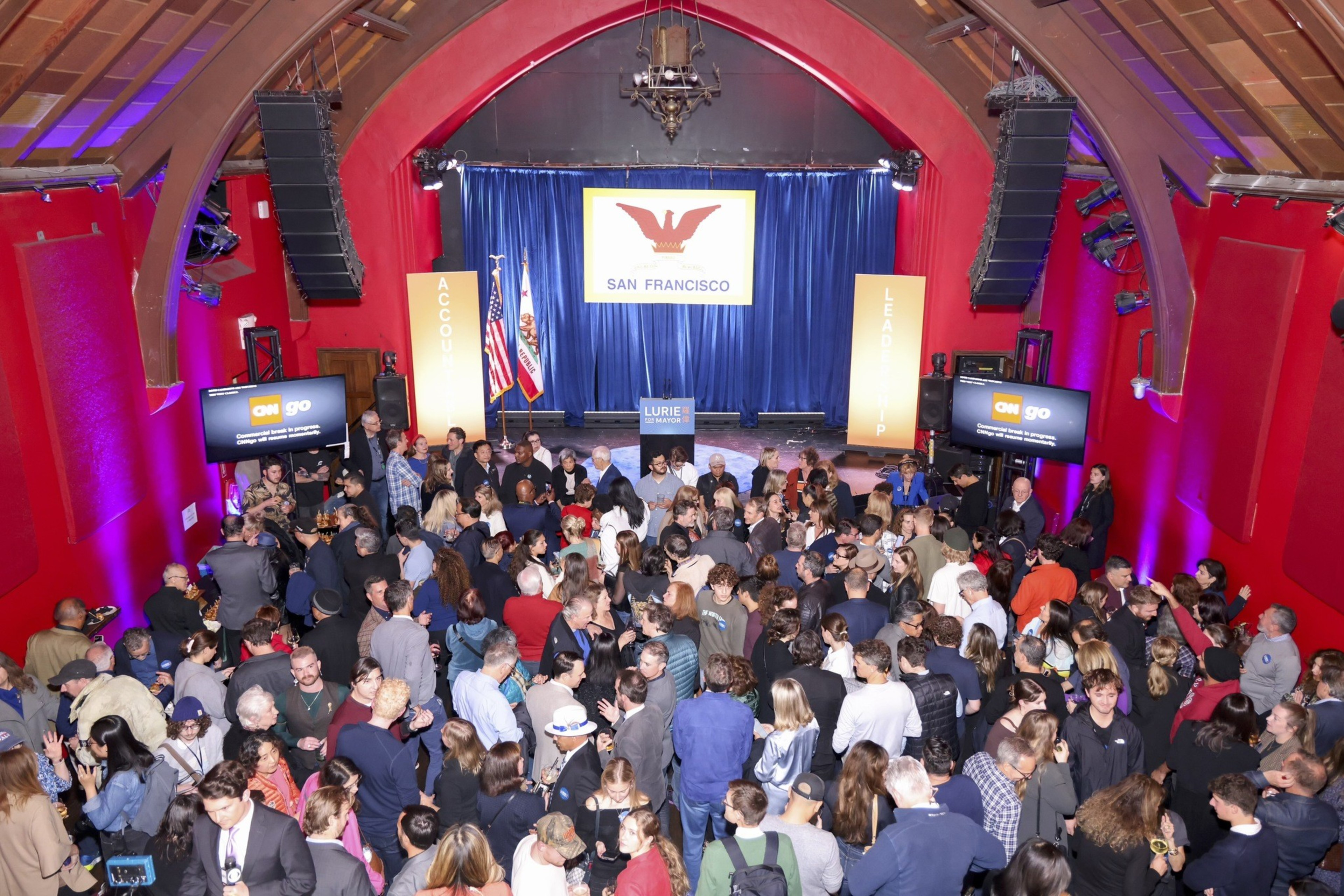 A large crowd gathers in an auditorium with red walls, watching a stage displaying &quot;San Francisco&quot; with two flags and a sign reading &quot;Lurie for Mayor.&quot;