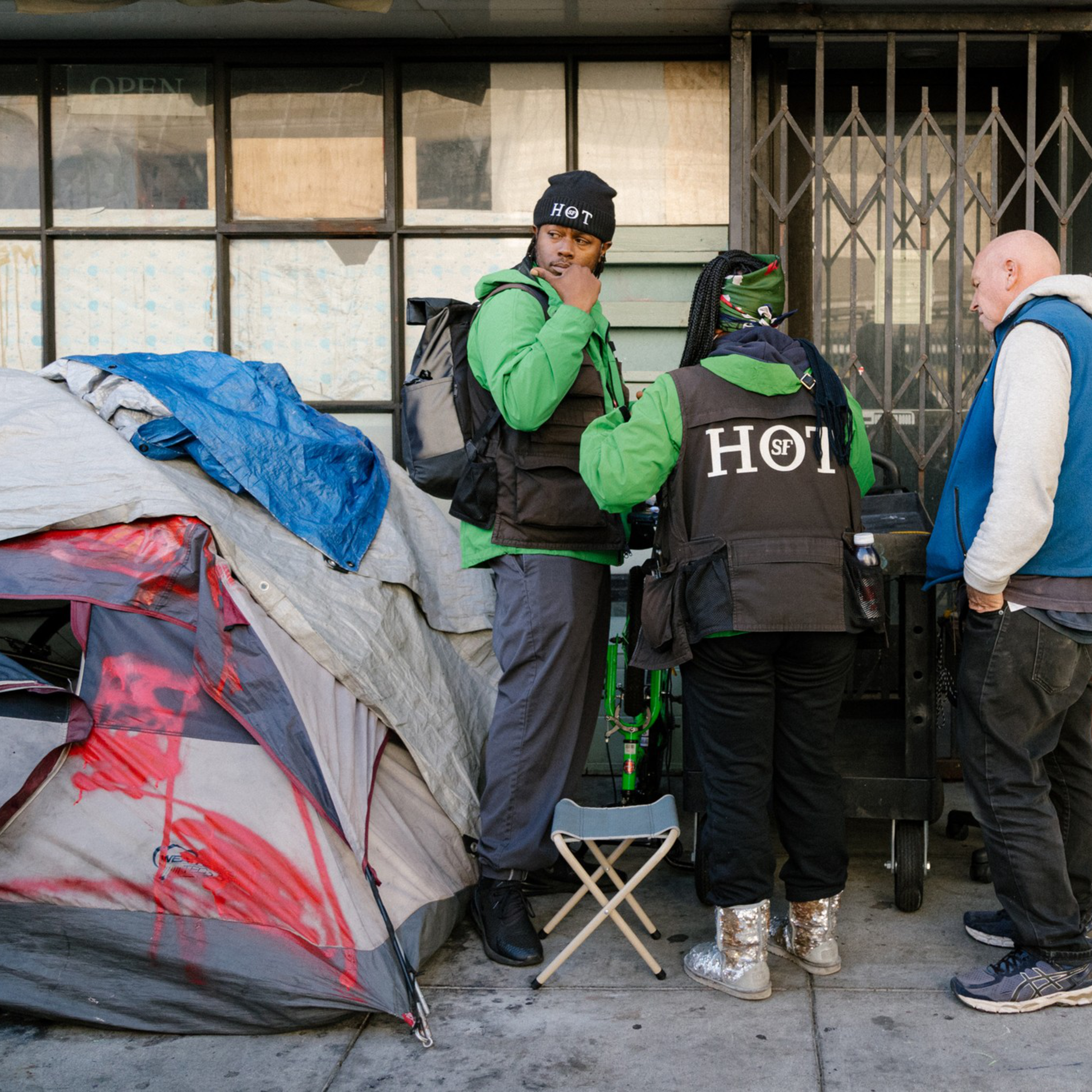 Four people are gathered on a sidewalk near a makeshift shelter. Two wear green and black "HOT" jackets, one wears an orange vest, and another wears a blue vest.