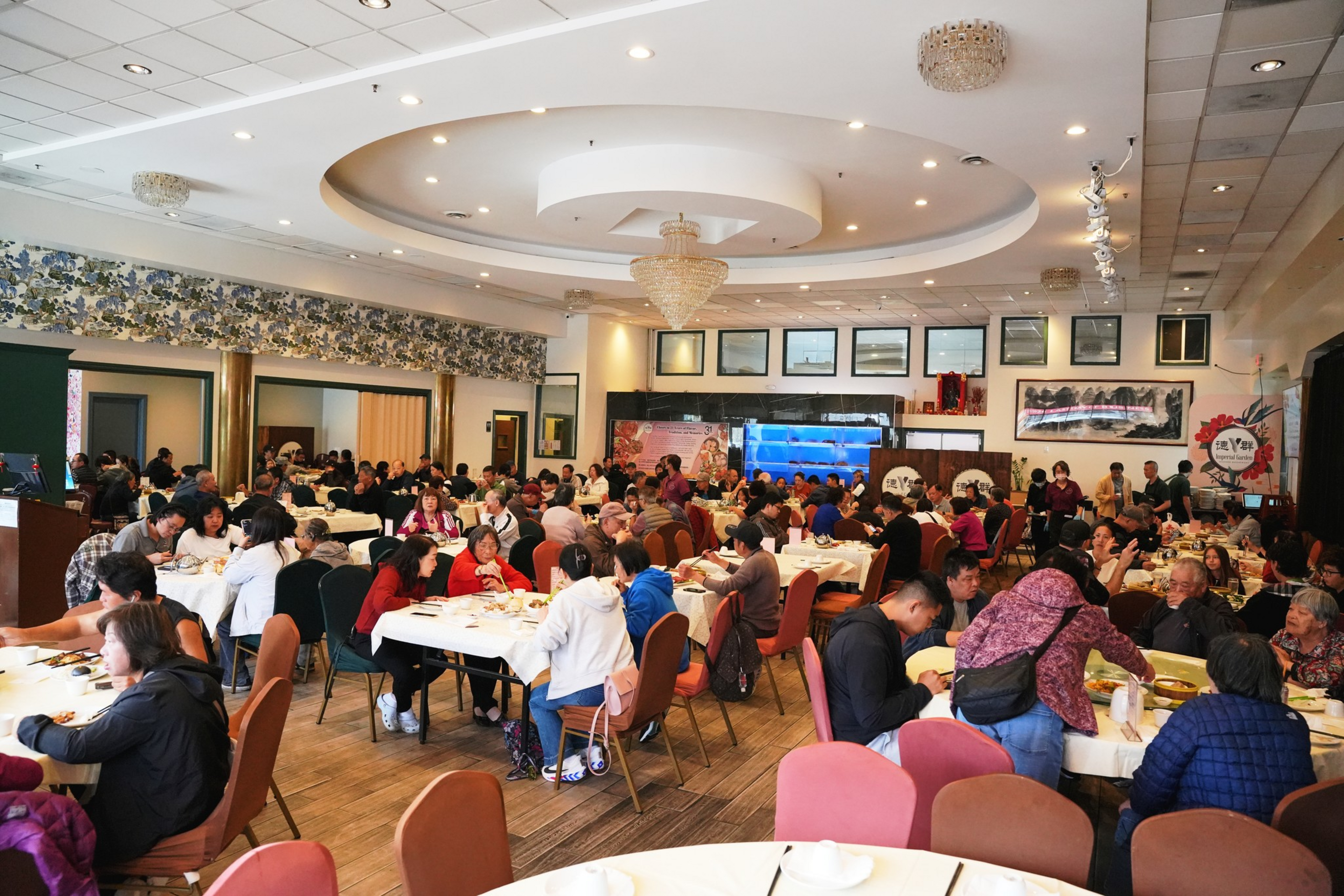 A large dining hall with many people seated at round and rectangular tables, eating and socializing under a circular ceiling light fixture.