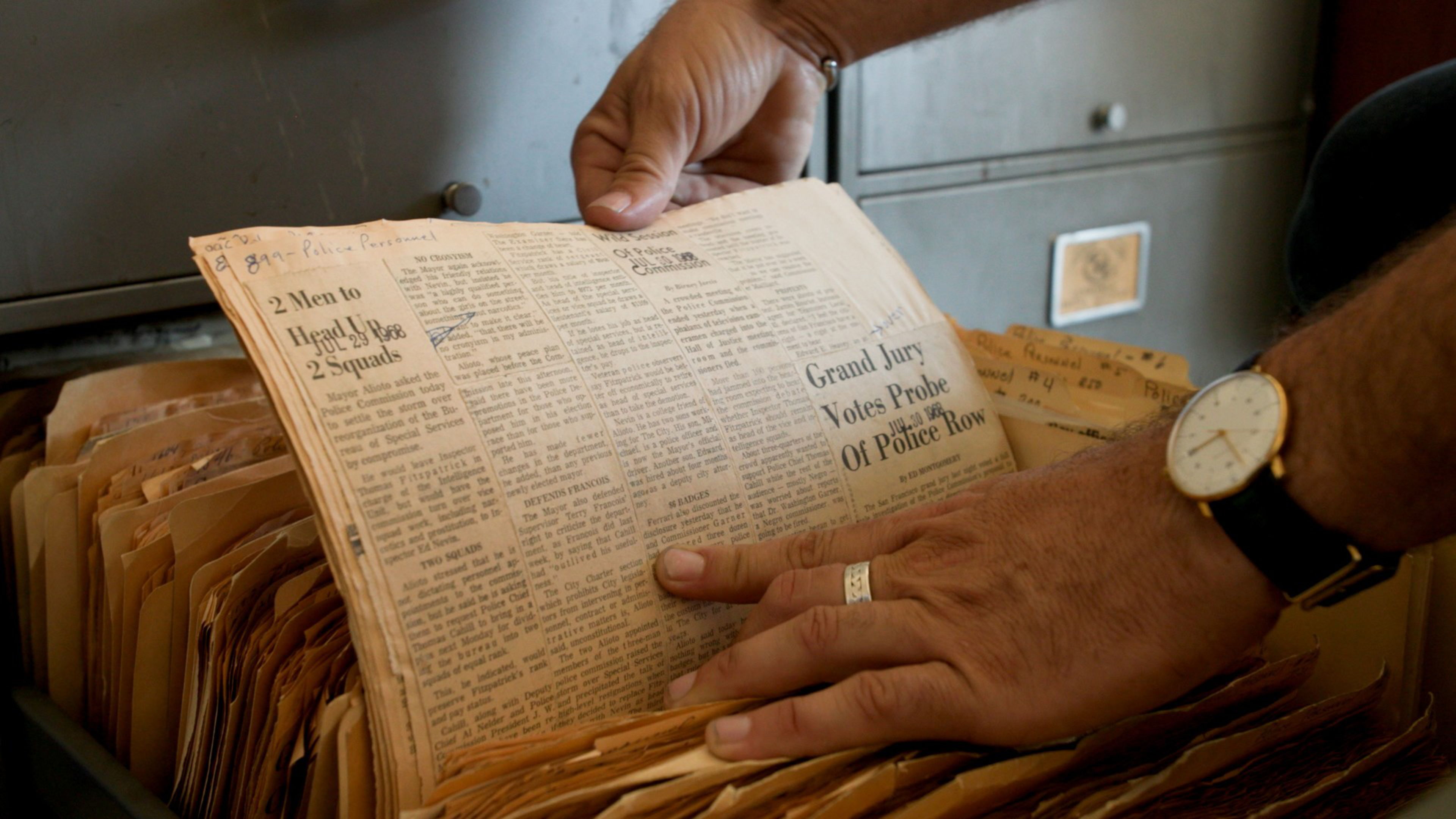 Hands with a watch and ring hold and flip through old newspaper clippings stored in a filing cabinet drawer filled with manila folders.