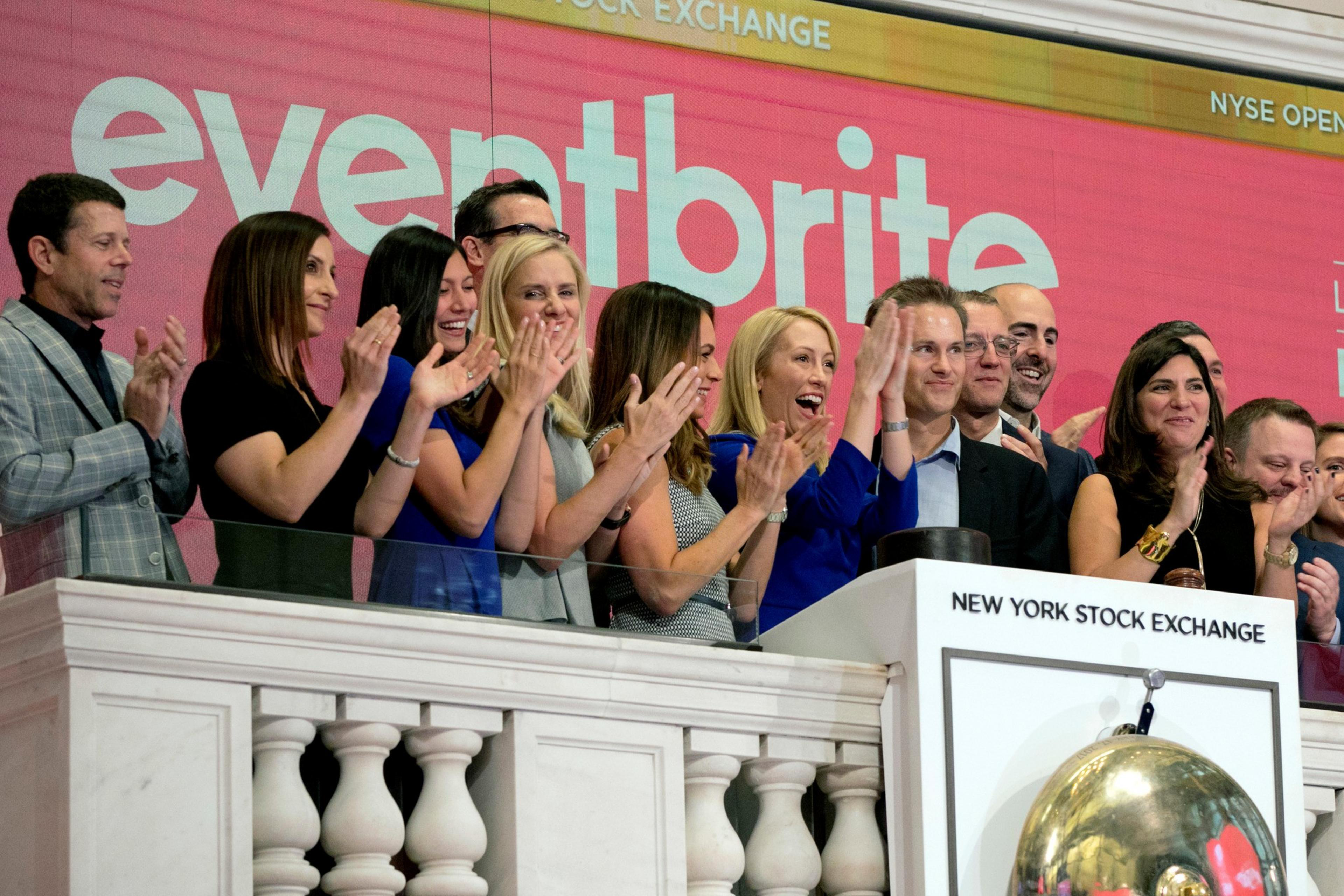 A group of people stand together, clapping and smiling, in front of a large "Eventbrite" sign at the New York Stock Exchange balcony.