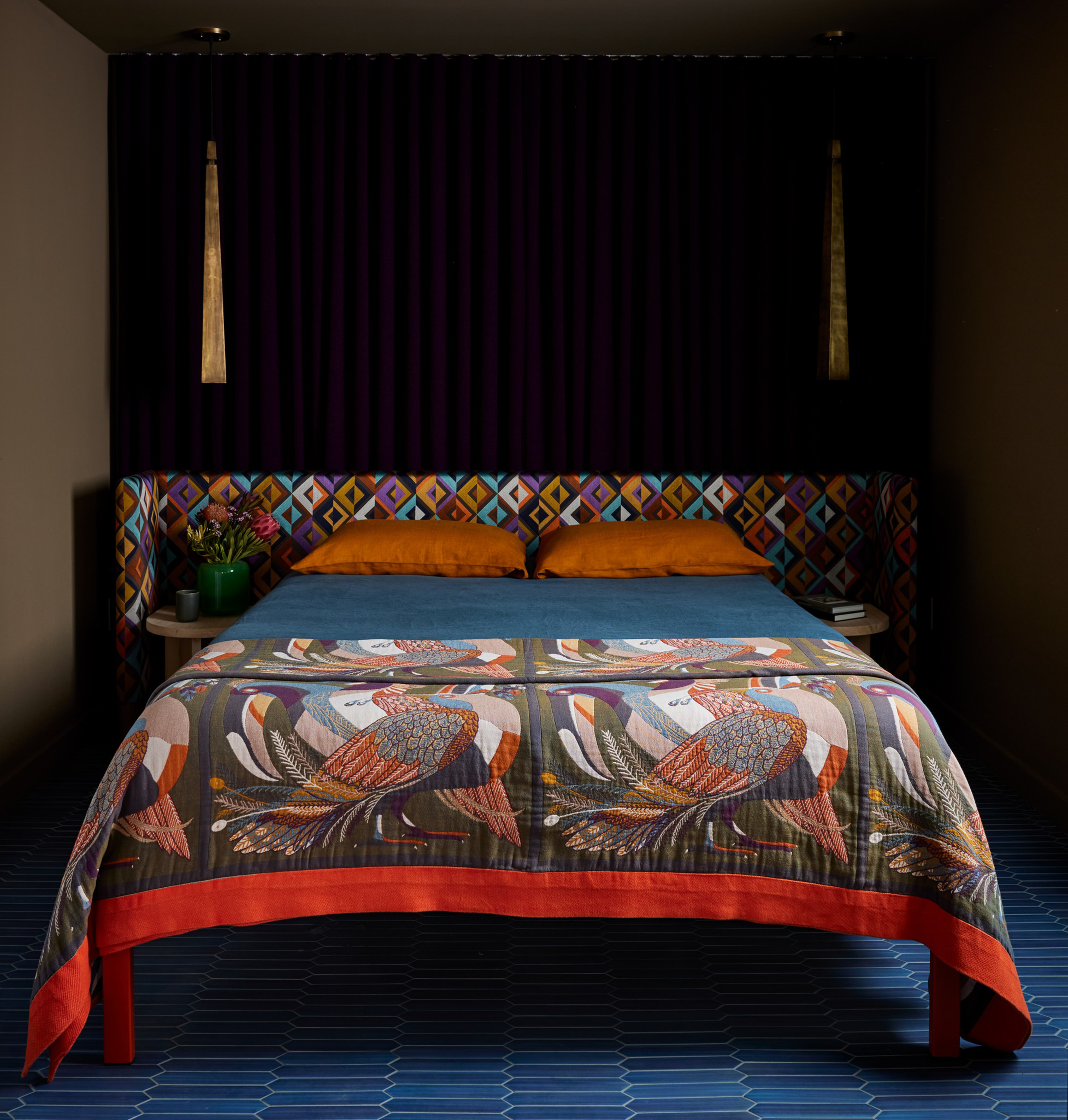 A vibrant bedroom with a geometric-patterned headboard, orange pillows, and a colorful bird-themed bedspread. Two pendant lights and a bedside table with flowers complete the scene.