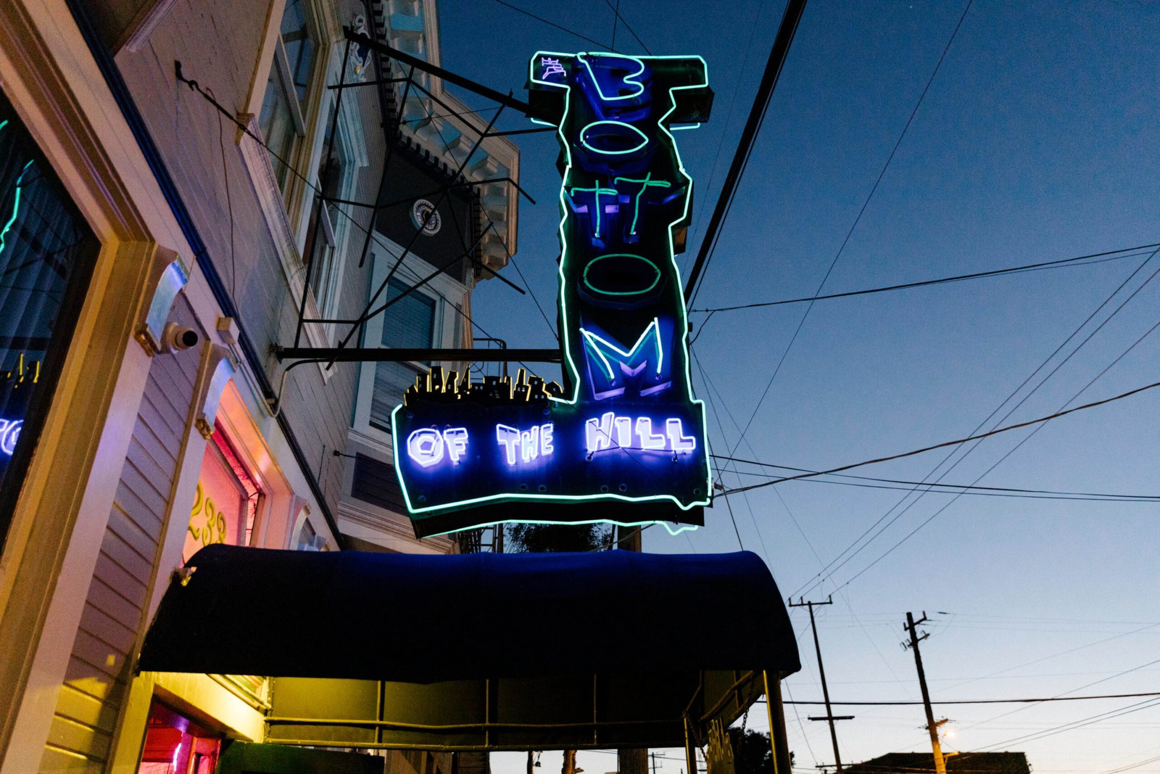 A neon sign reading "BOTTOM OF THE HILL" glows in blue and green above a storefront at dusk with power lines and a clear sky in the background.