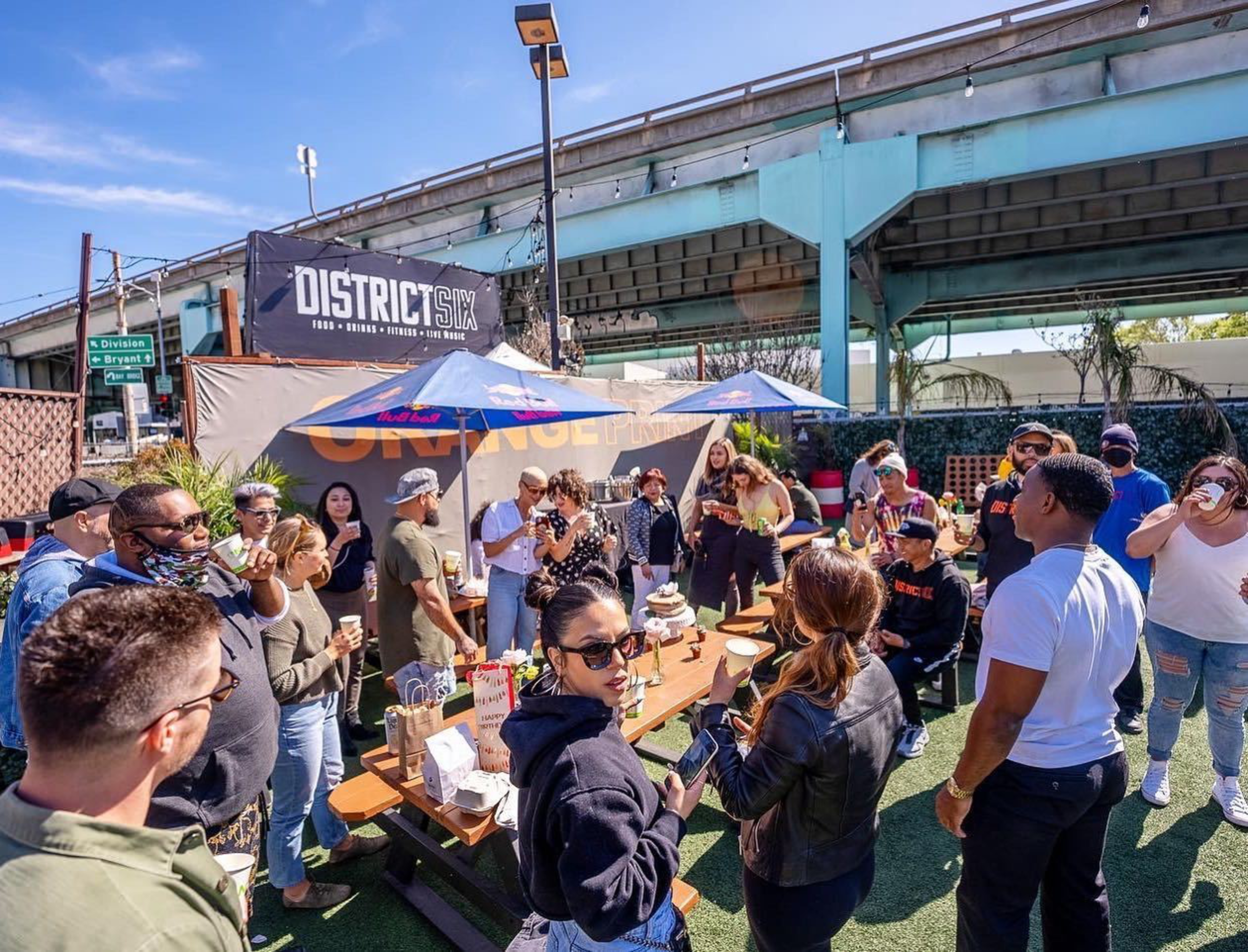 People gather outdoors under umbrellas and a highway overpass, socializing around picnic tables with drinks and snacks in a bright, sunny setting.