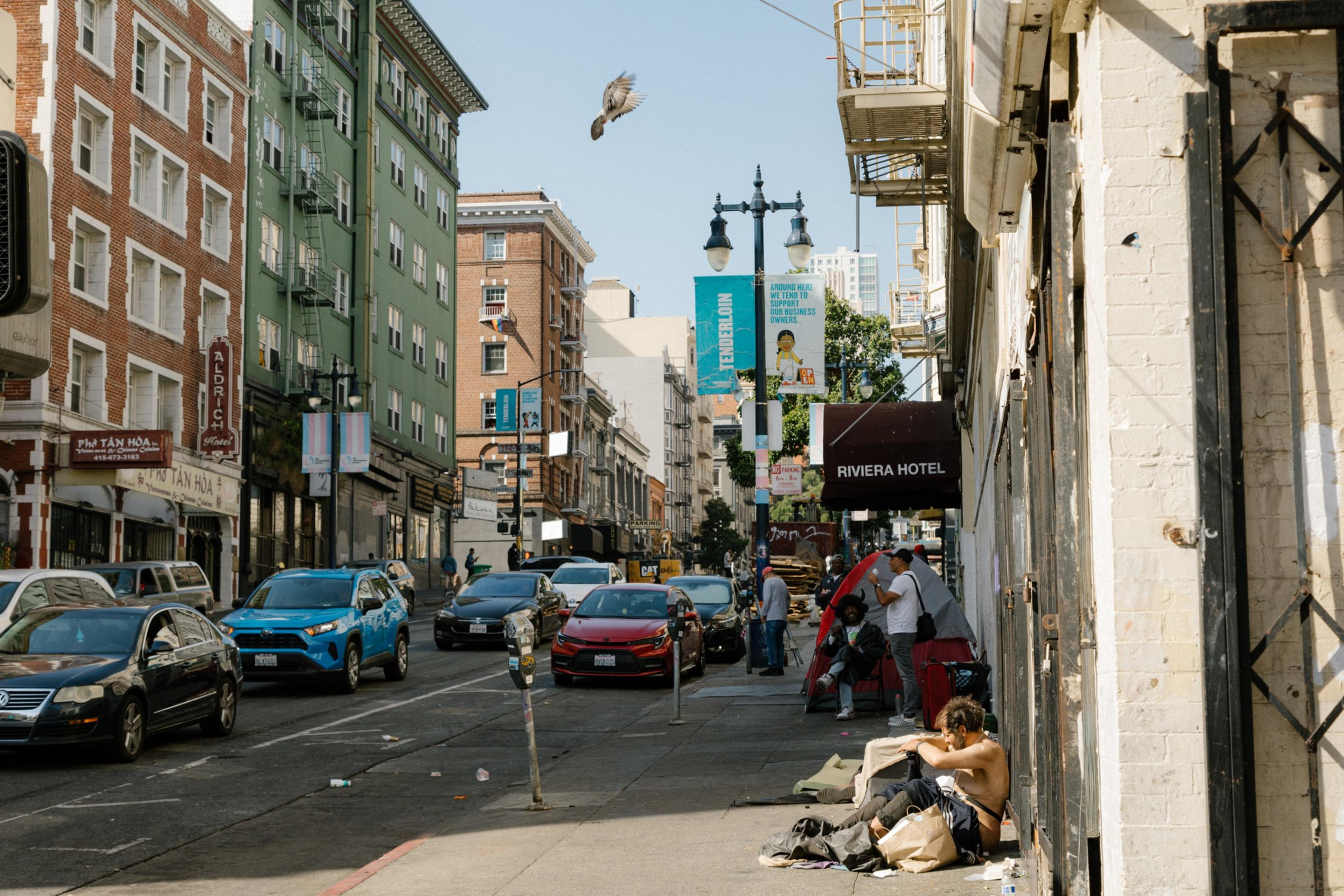 A shirtless man sits on the sidewalk of a busy street