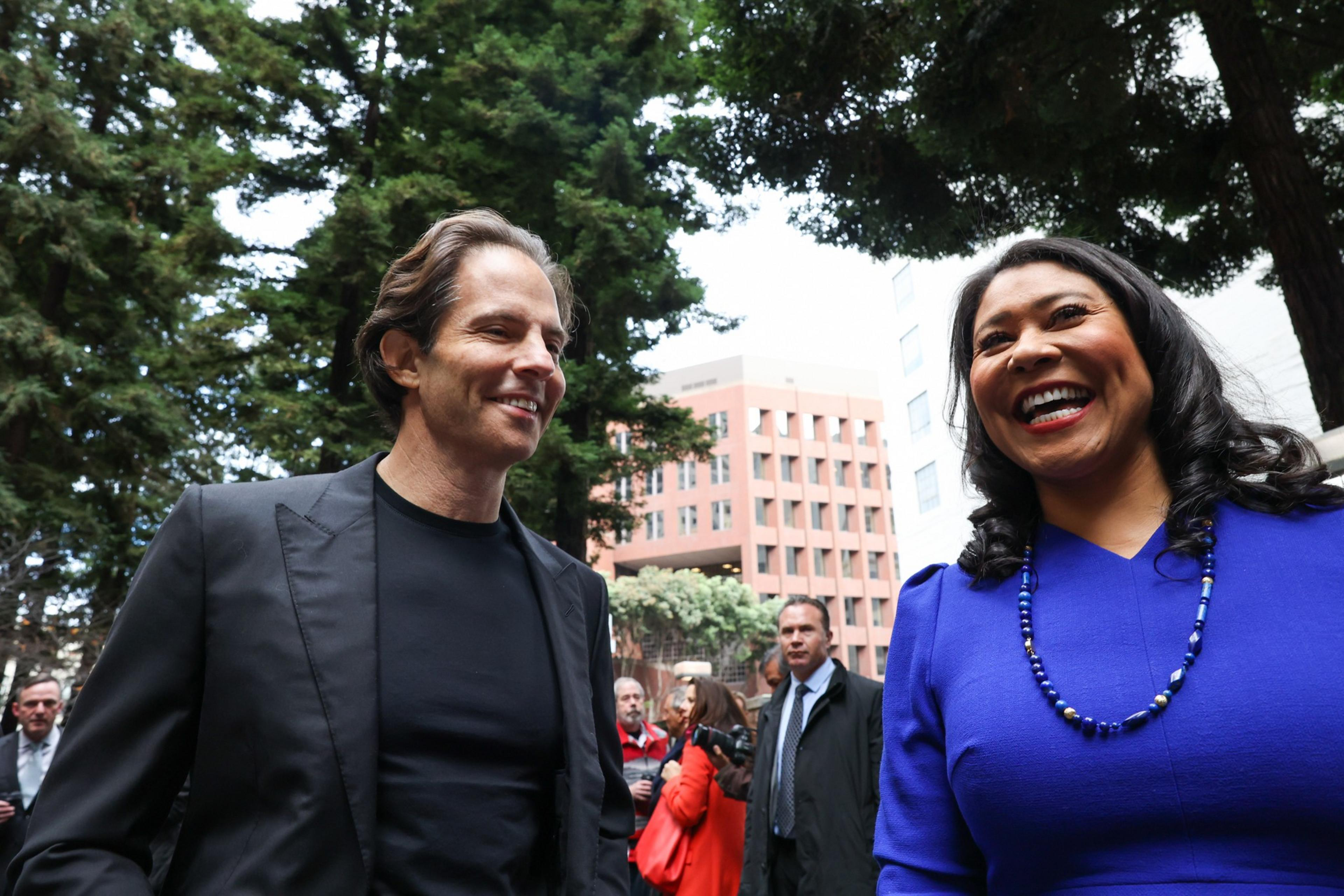 A man in a black suit and a woman in a blue dress with a beaded necklace are smiling and talking outdoors, with several people and trees in the background.