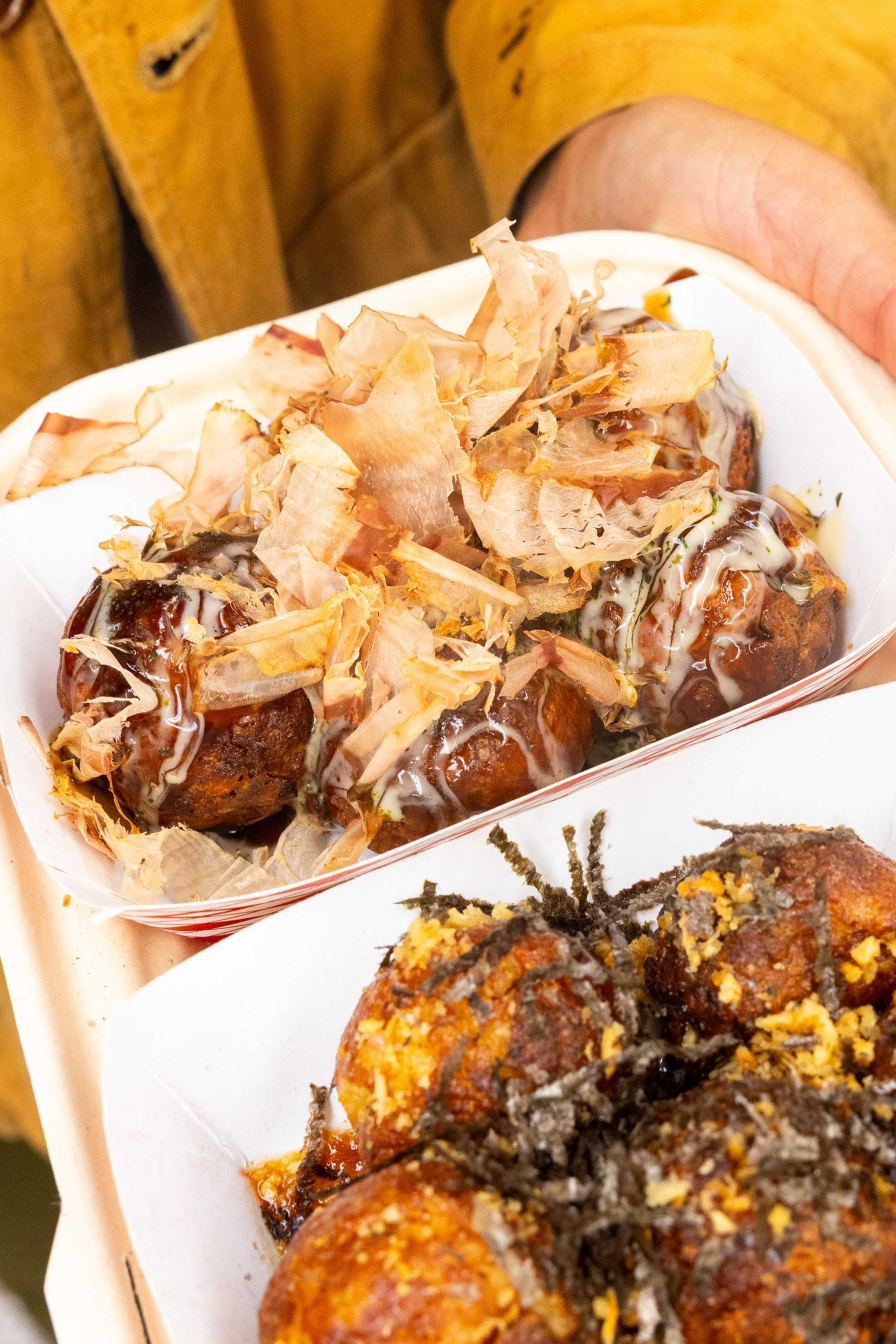 Two trays of takoyaki balls topped with sauce, mayo, dried bonito flakes on one tray, and crispy seaweed flakes on the other tray.