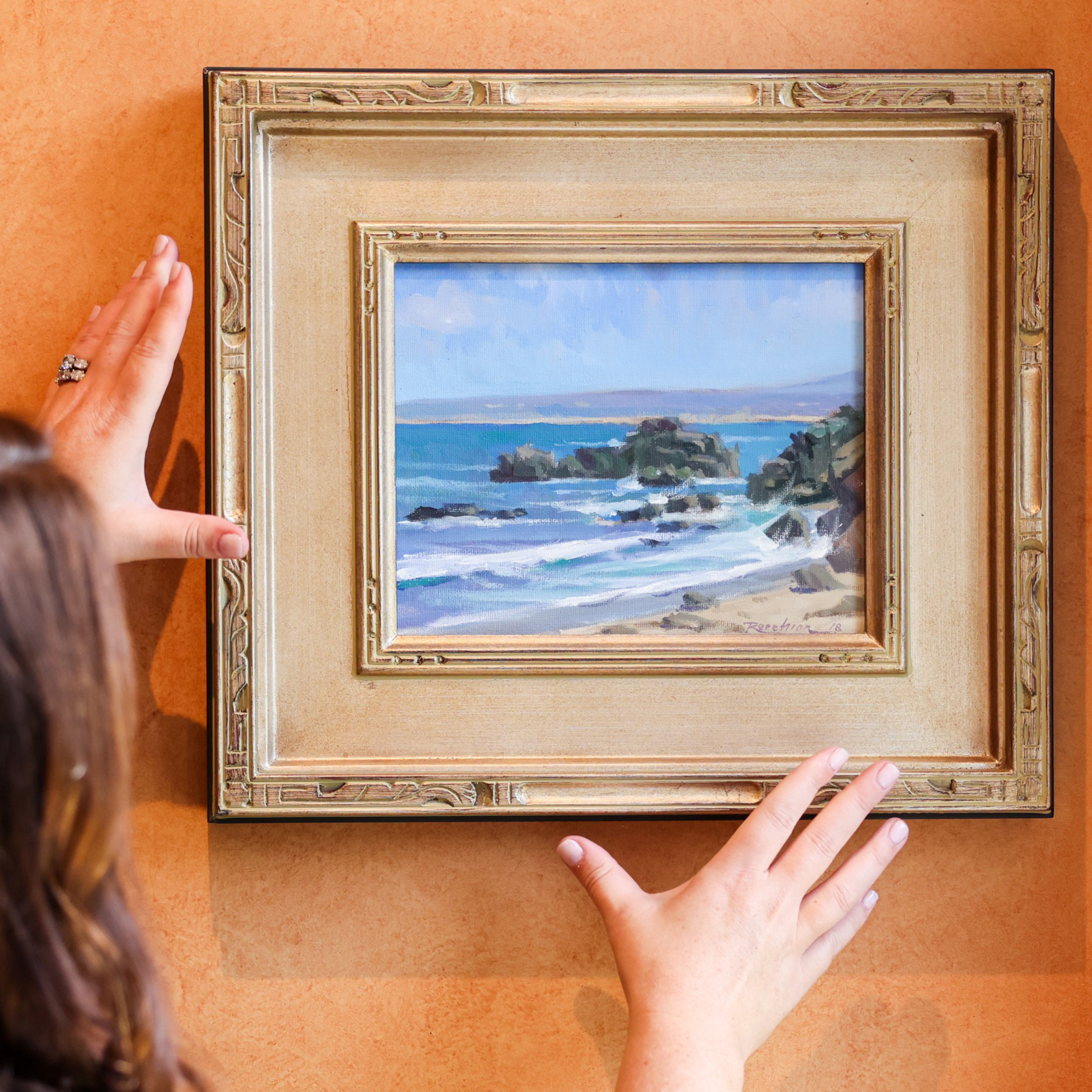 A person is adjusting a framed painting of a coastal scene, featuring blue waves and rocky shores, against a warm-toned wall.