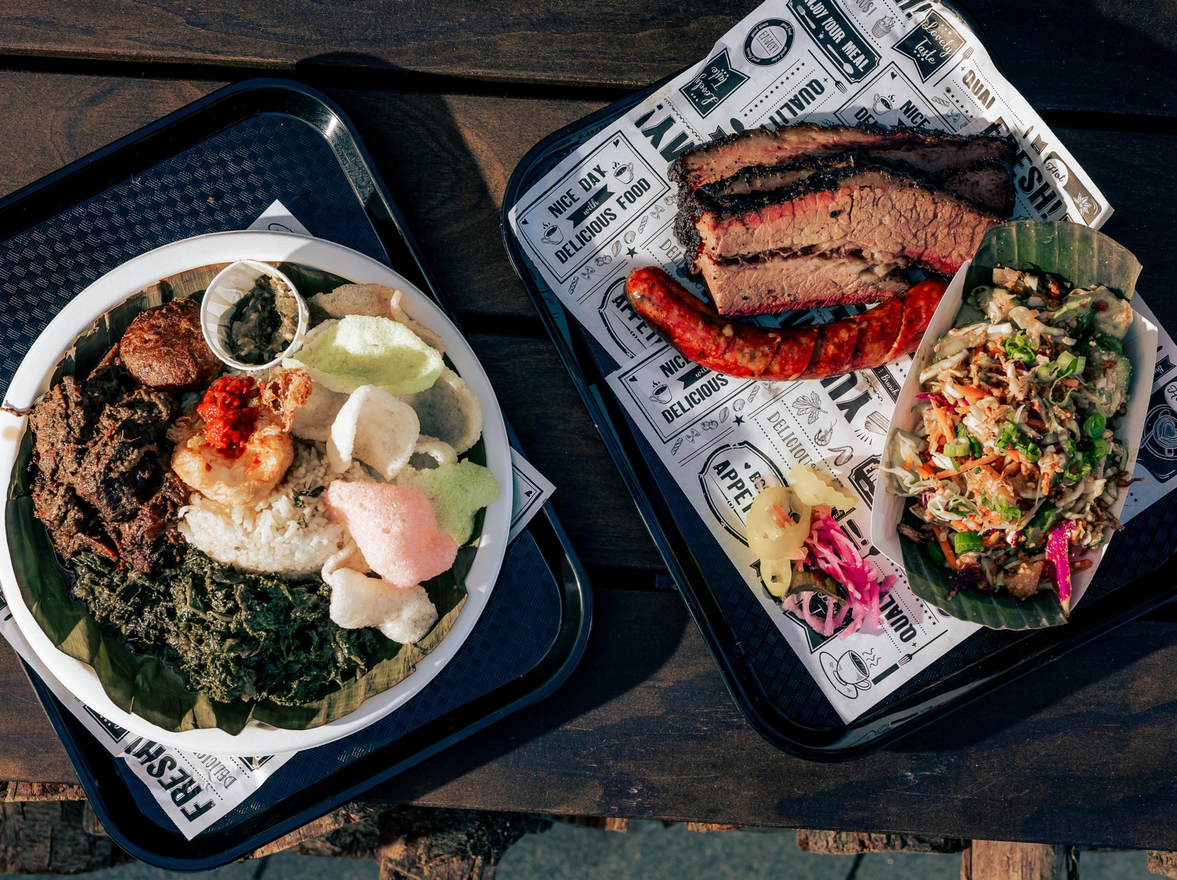 The image shows two trays of food: one with rice, meat, greens, crackers, and sauce, and the other with sliced brisket, sausage, pickles, and a vegetable salad.