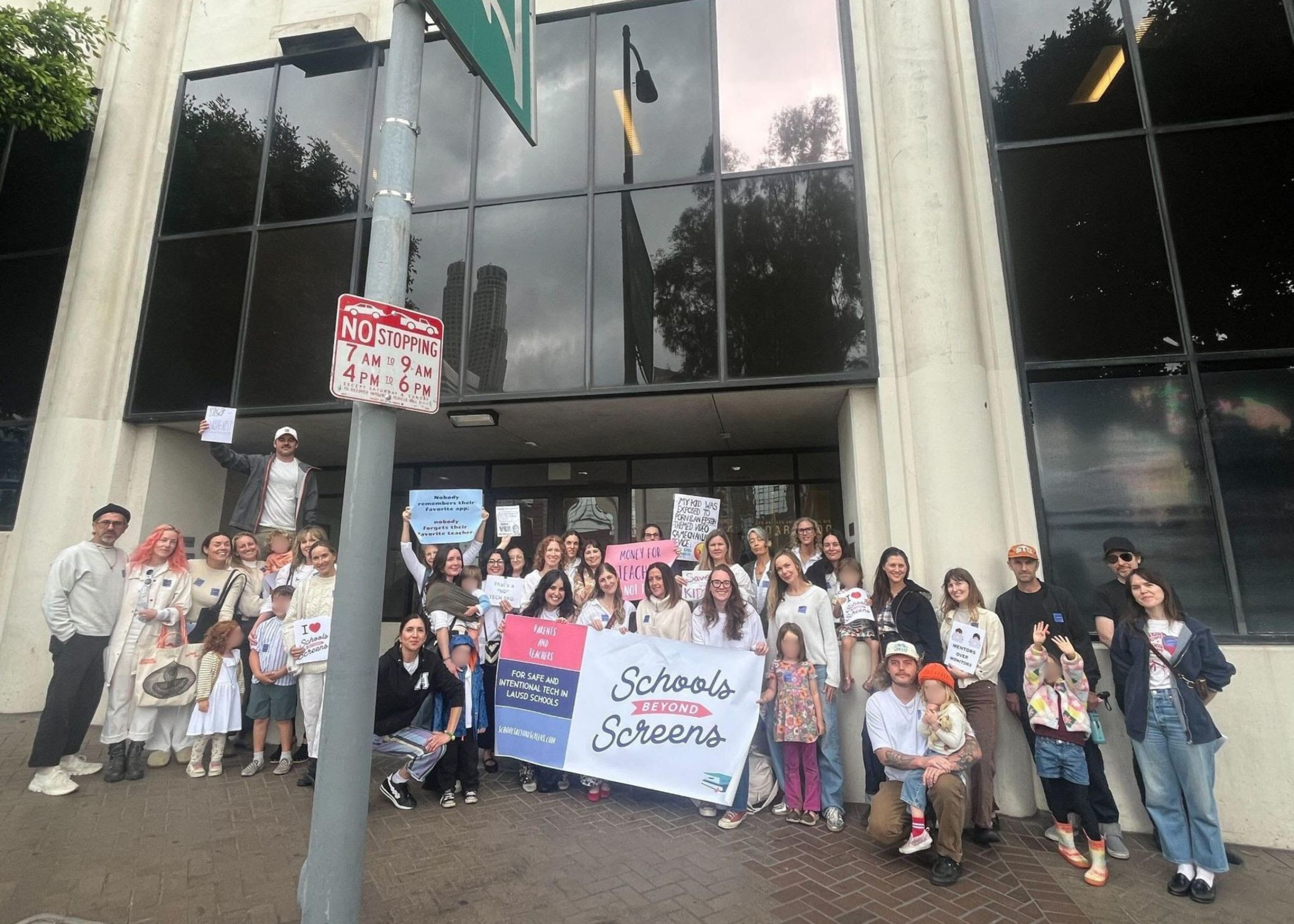 A diverse group of people, including adults and children, stand outside a building holding signs and a large “Schools Beyond Screens” banner.