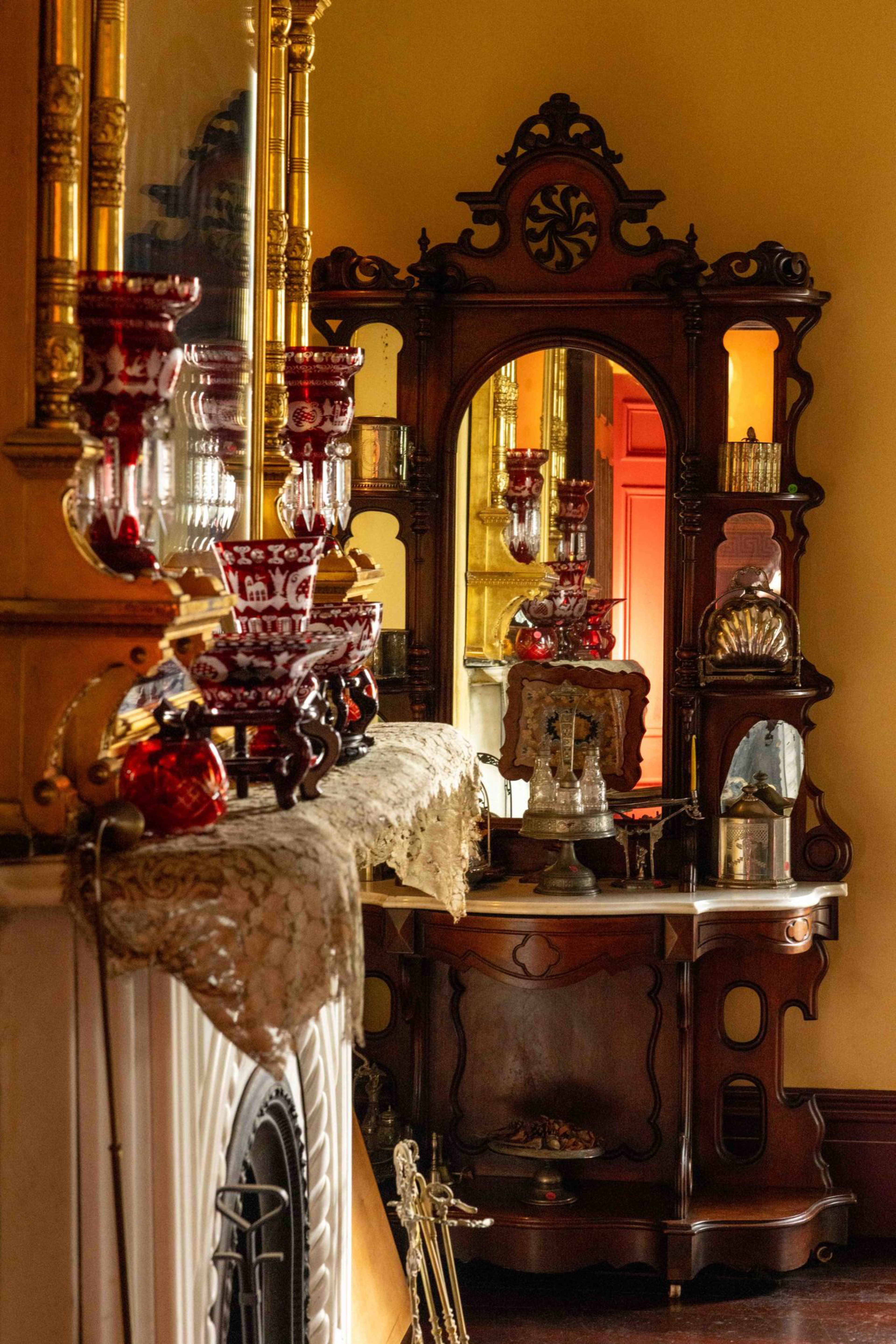 Ornate wooden mantel and vanity mirror hold red glassware, lace cloth, and vintage decor in a warmly lit, antique-style room.