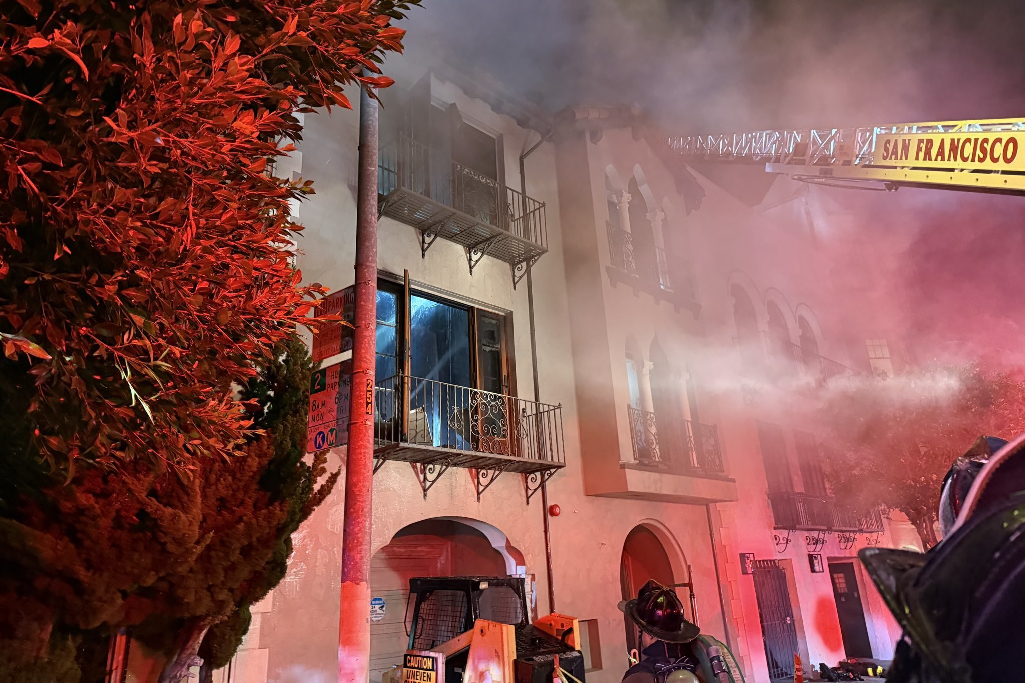 Firefighters spray water from a ladder truck labeled "San Francisco" onto a smoky building at night. Bright red lights illuminate nearby trees and walls.
