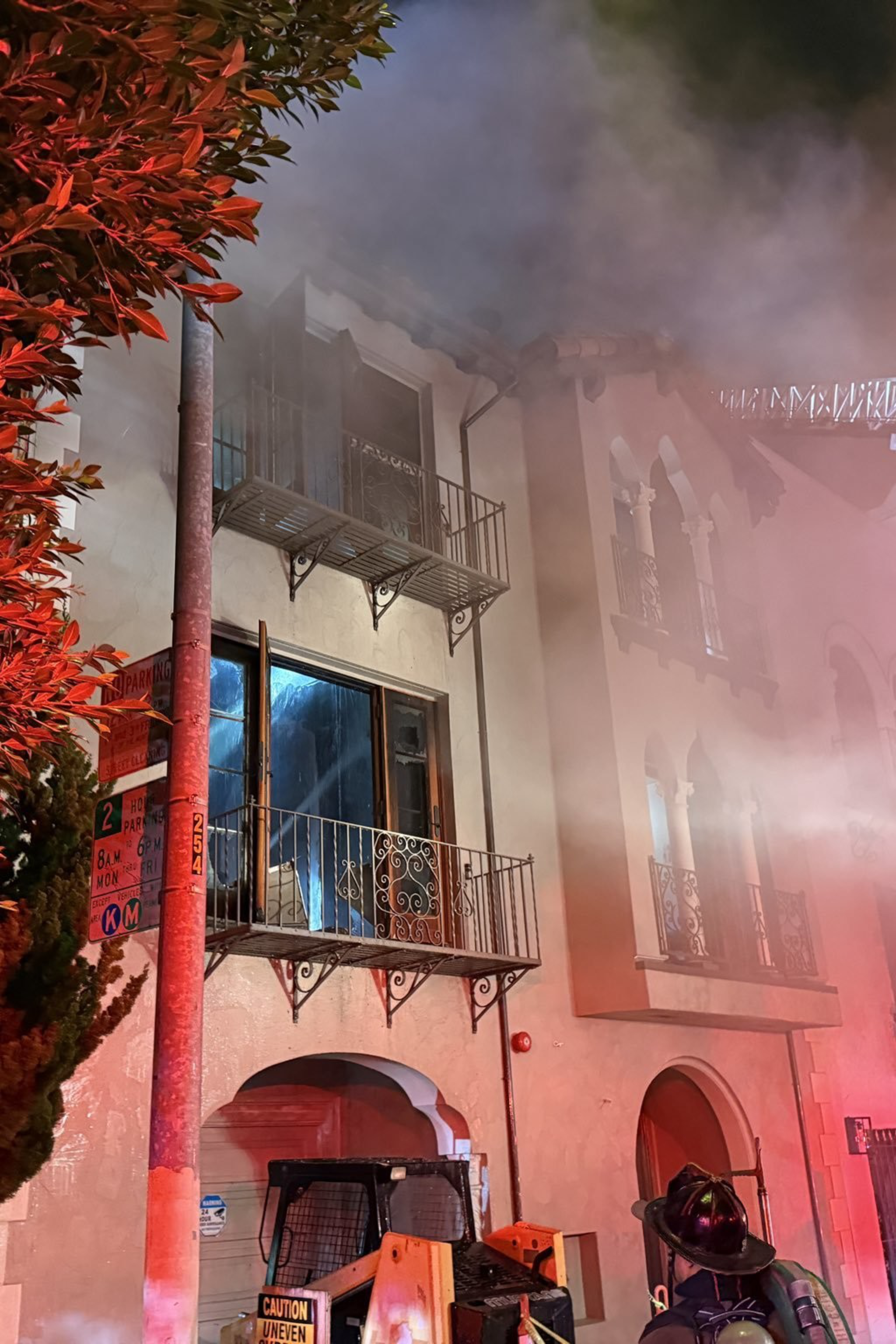 Firefighters spray water from a ladder truck labeled "San Francisco" onto a smoky building at night. Bright red lights illuminate nearby trees and walls.