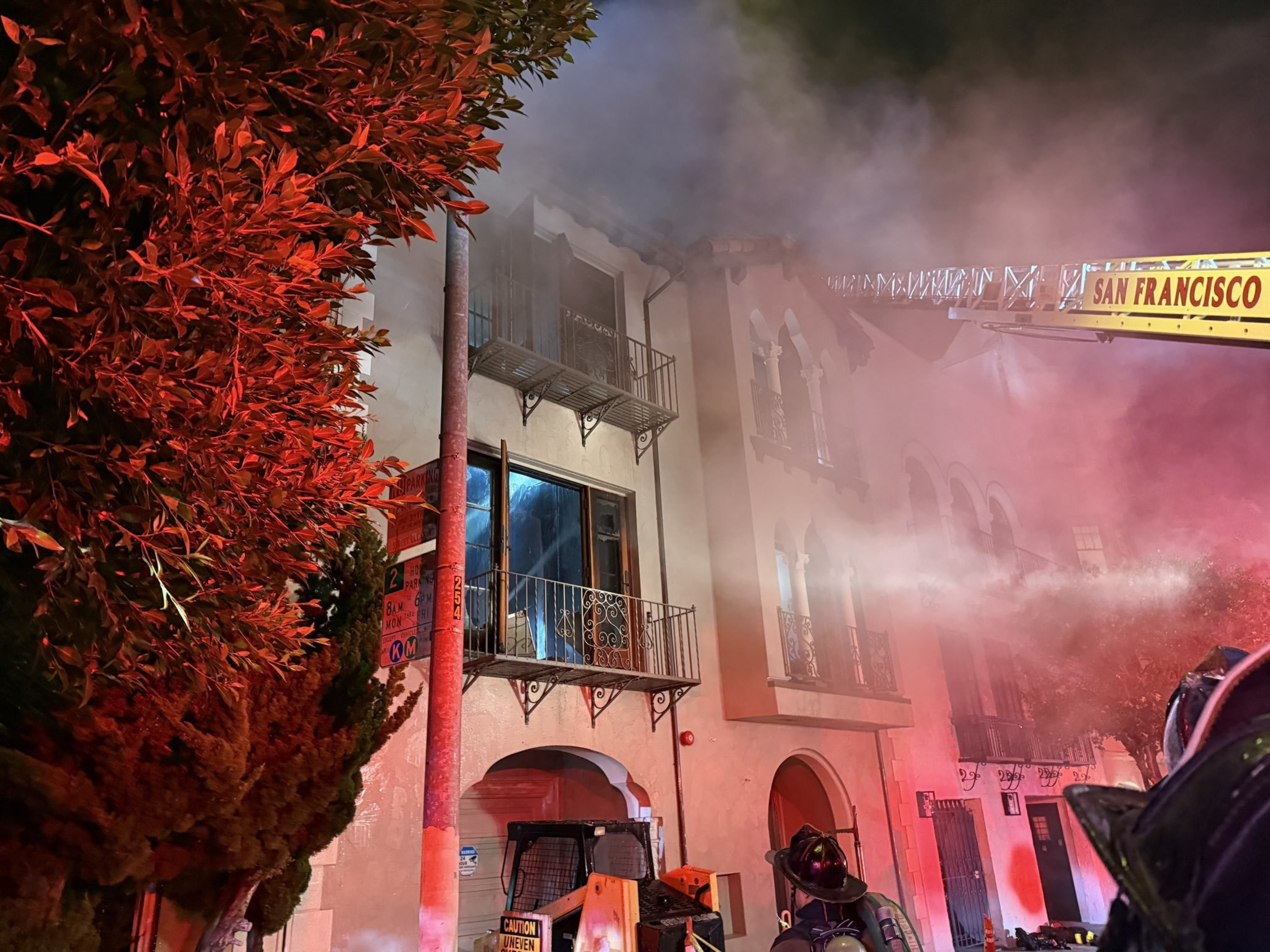 Firefighters spray water from a ladder truck labeled "San Francisco" onto a smoky building at night. Bright red lights illuminate nearby trees and walls.