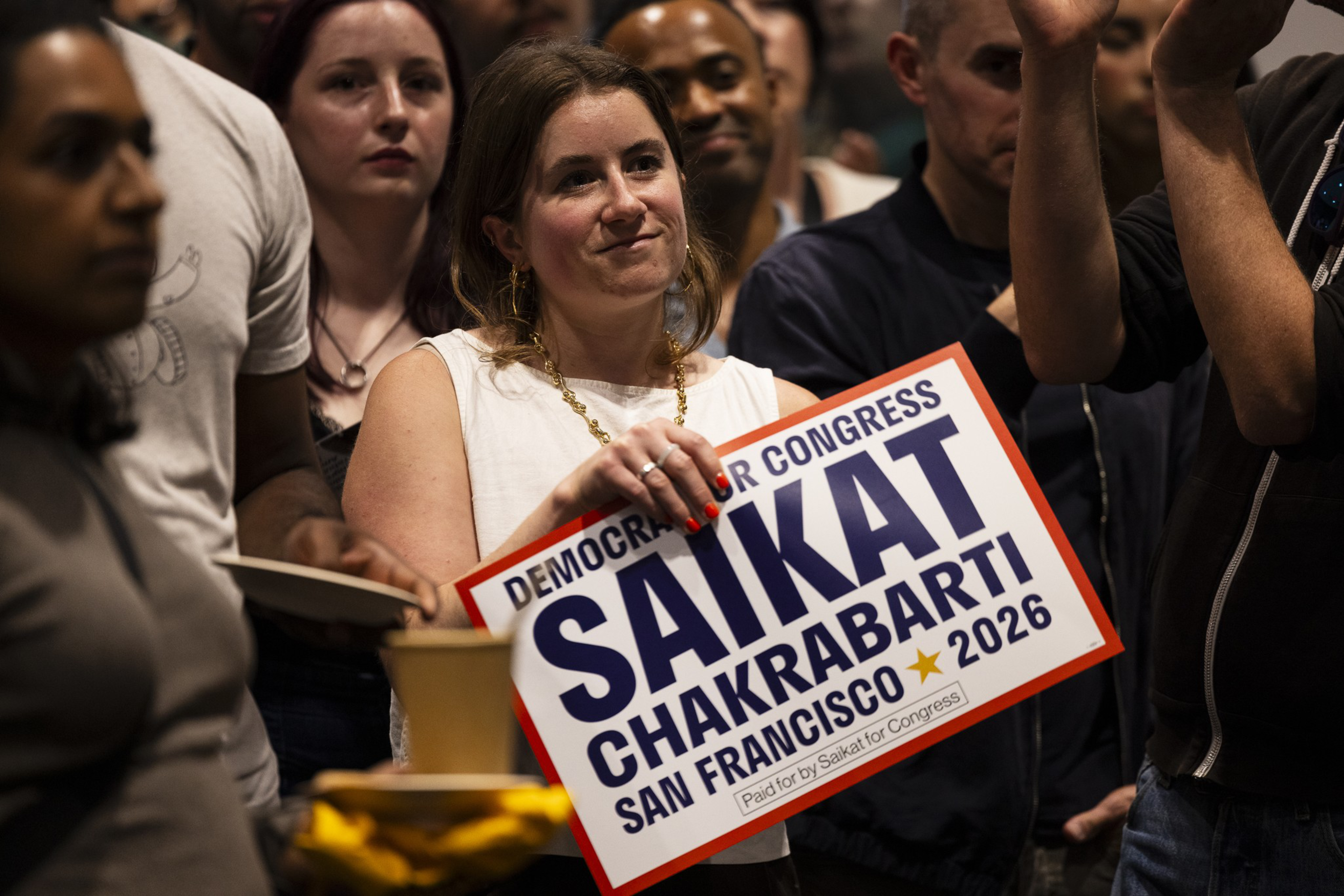 A woman in a white top holds a political sign for Saikat Chakrabarti’s 2026 San Francisco Democratic Congress campaign, surrounded by a diverse crowd.
