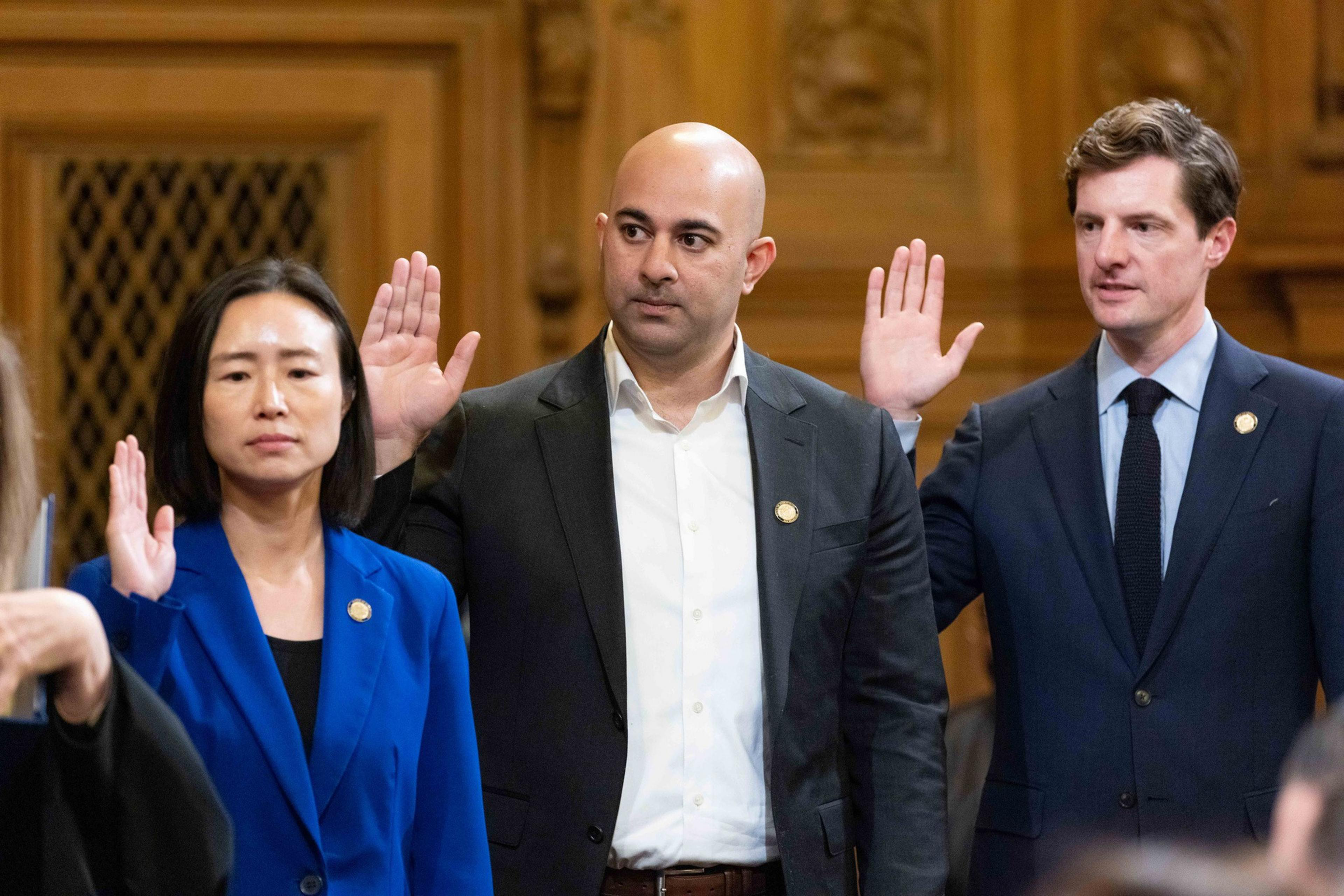 Three individuals are raising their right hands, likely taking an oath. They wear formal attire, with two men in suits and a woman in a blue blazer.
