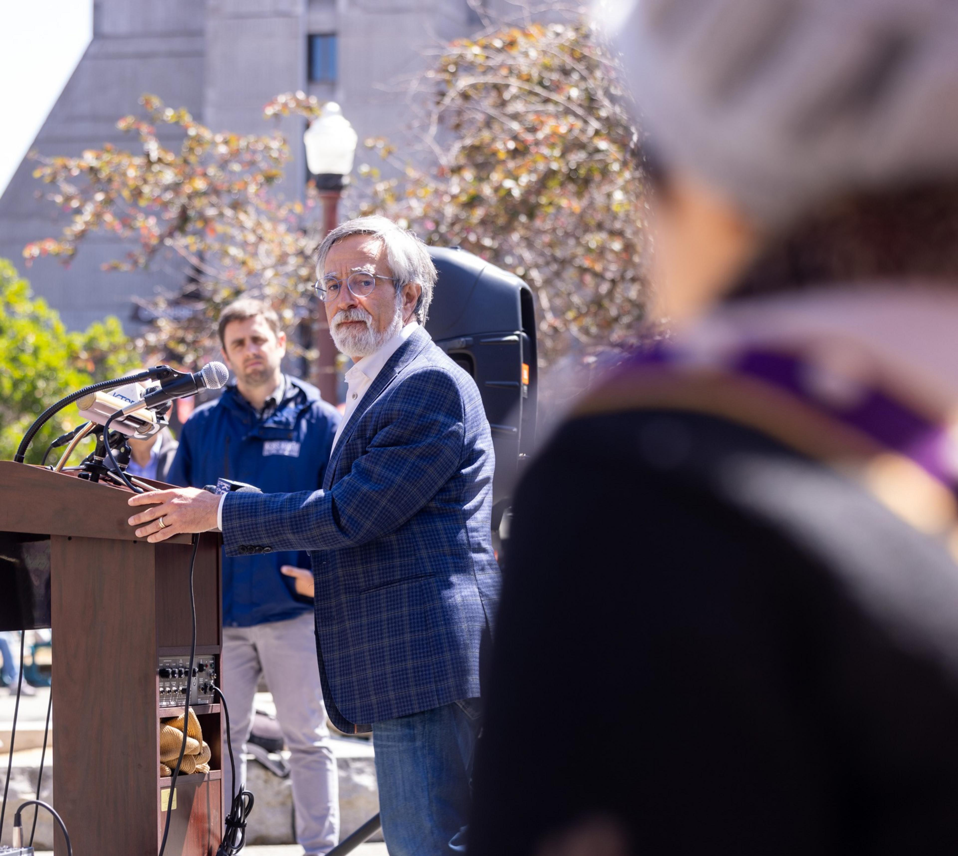 An older man with gray hair and glasses speaks at an outdoor podium. A man in the background watches, and a blurred figure is in the foreground.
