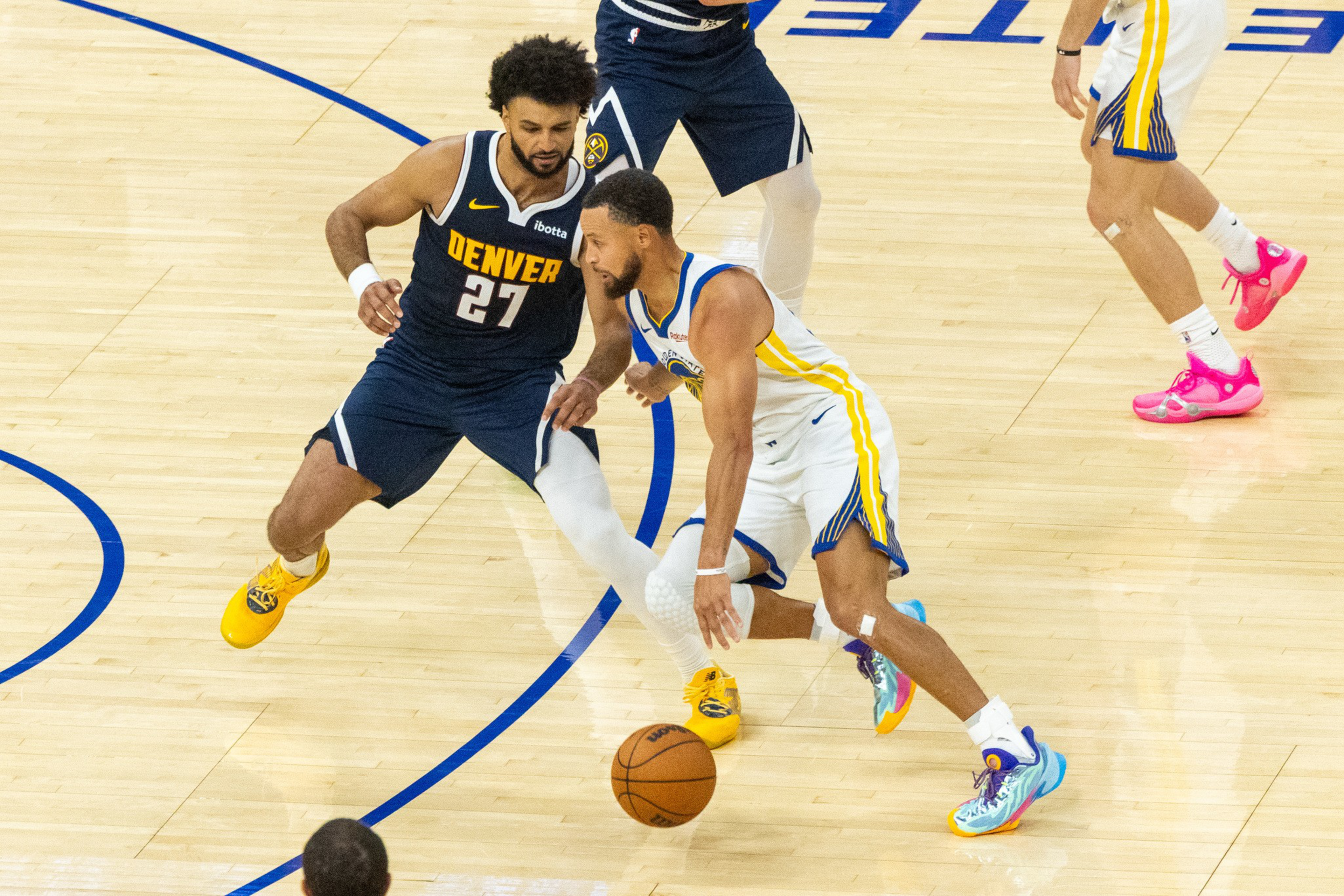 A Denver Nuggets player defends while a Golden State Warriors player dribbles a basketball on a hardwood court during a game.