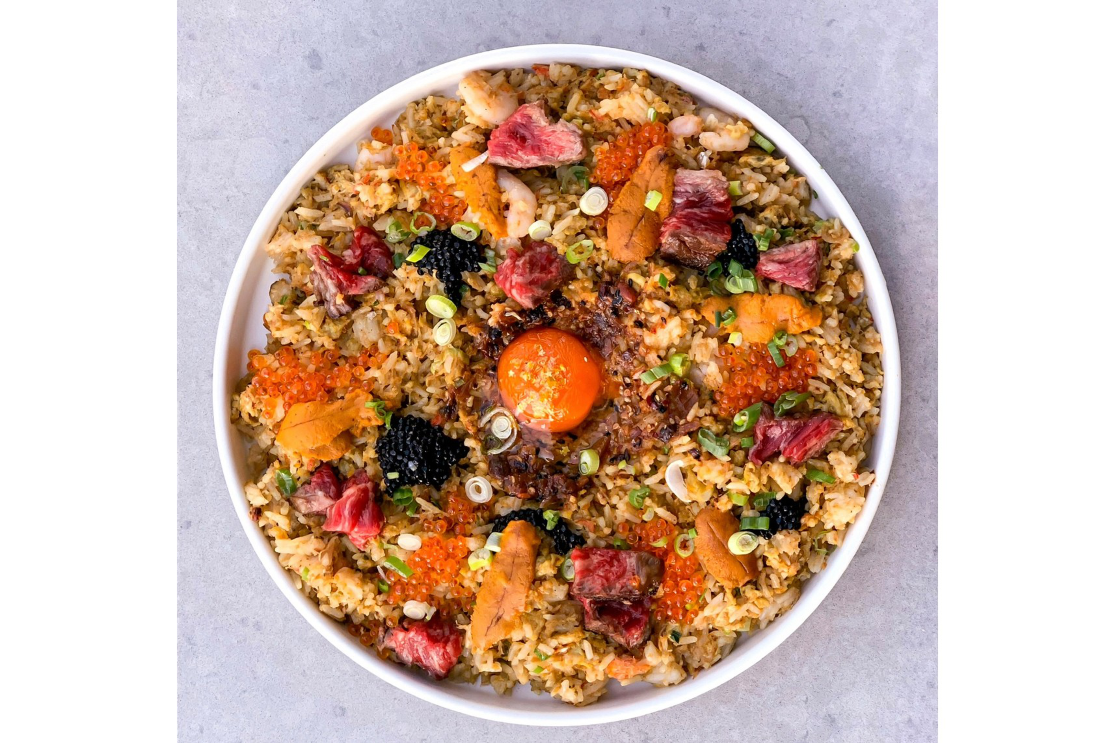 A bowl of fried rice topped with raw egg yolk, pieces of seared beef, shrimp, orange fish roe, black caviar, sea urchin, and chopped green onions.