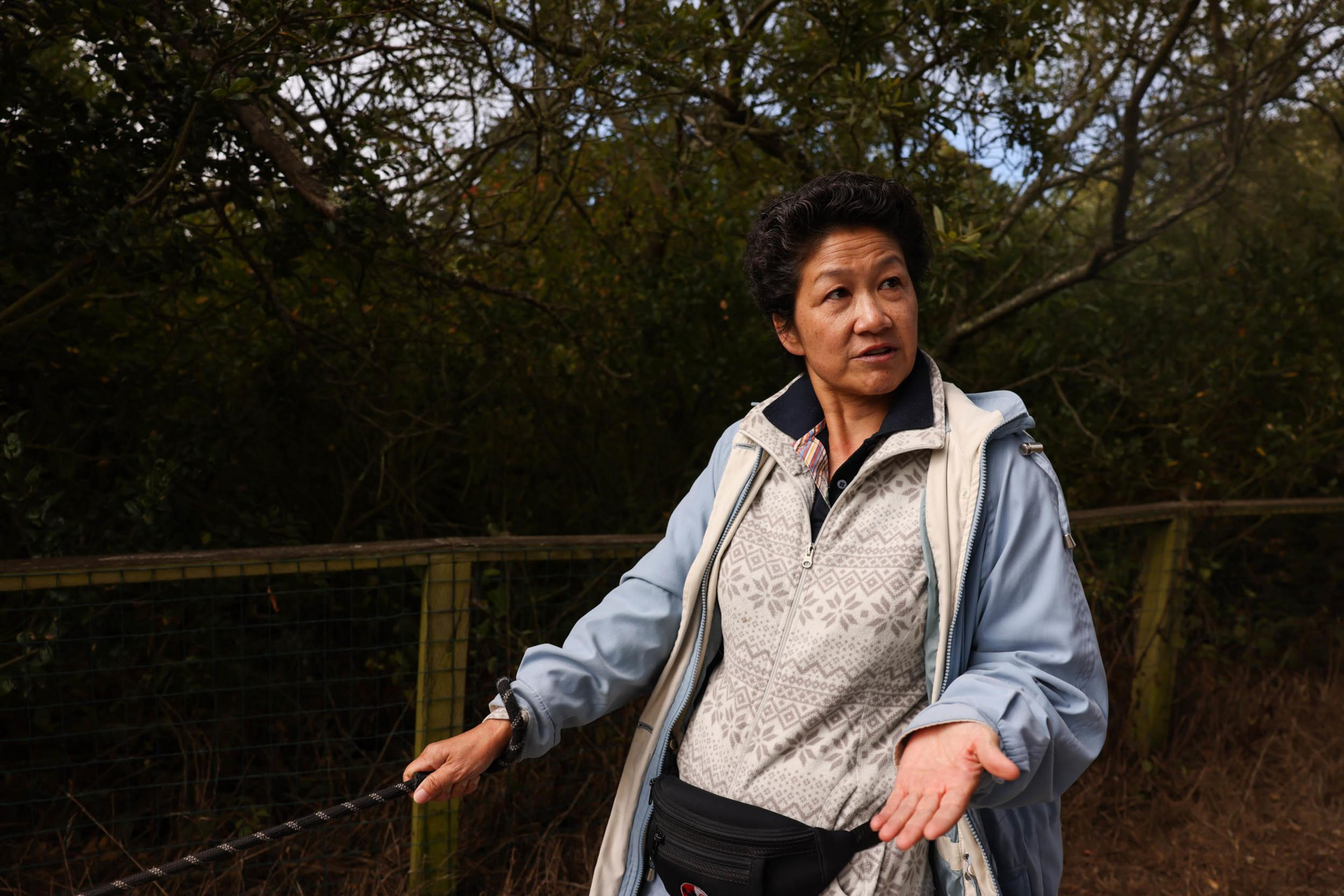 A person dressed in a light blue jacket and patterned sweater stands outdoors, gesturing with one hand while holding a leash in the other, with trees and a wooden fence behind them.