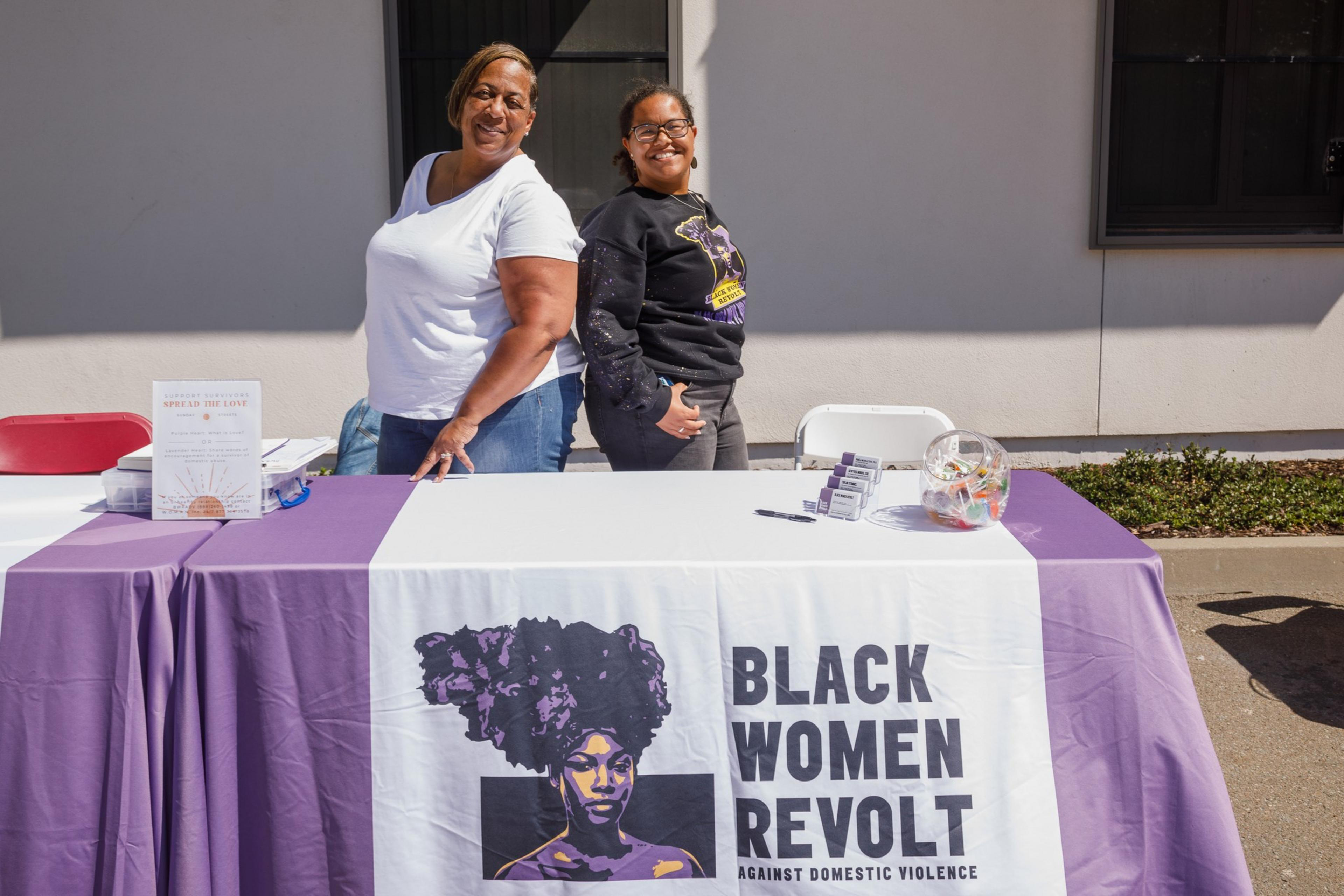 Two individuals stand behind a table with a banner reading &quot;BLACK WOMEN REVOLT AGAINST DOMESTIC VIOLENCE&quot;.