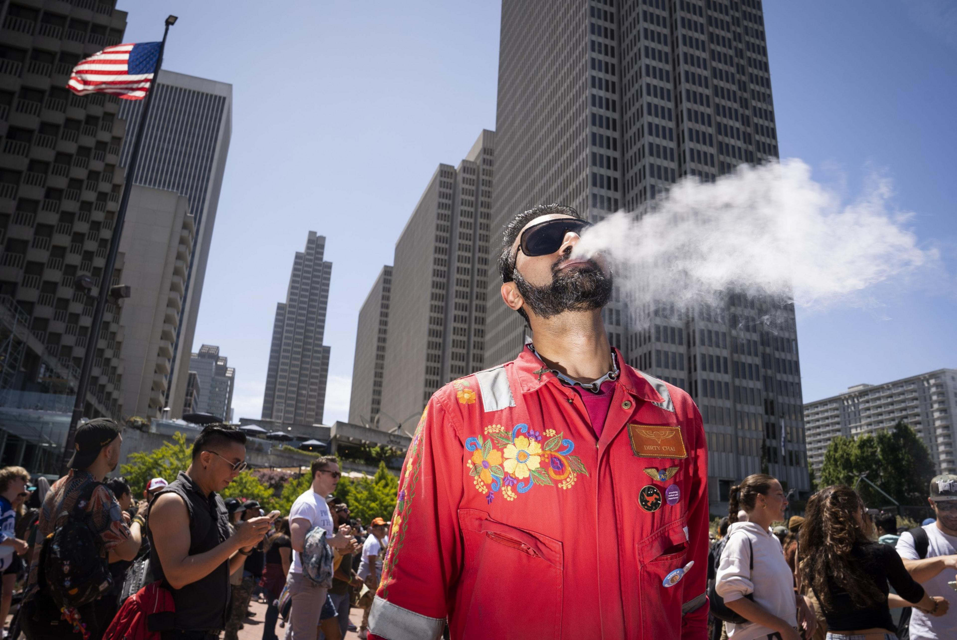 A person in a colorful red outfit blows vapor into the air, standing amidst a crowd in an urban setting with tall buildings and a U.S. flag nearby.