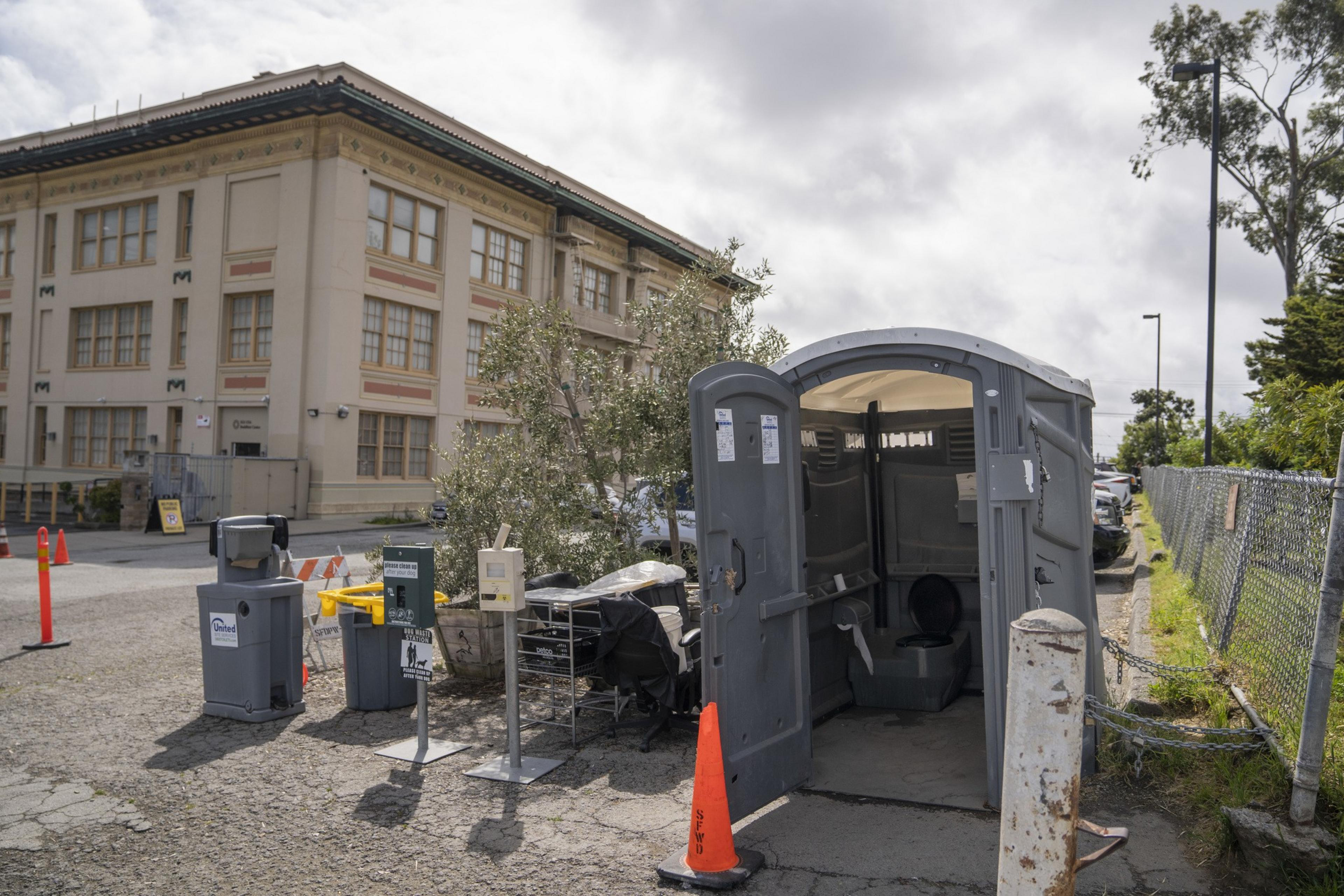 A portable toilet is open beside a beige building and a chain-link fence. Traffic cones, bins, and a small tree are nearby under a cloudy sky.