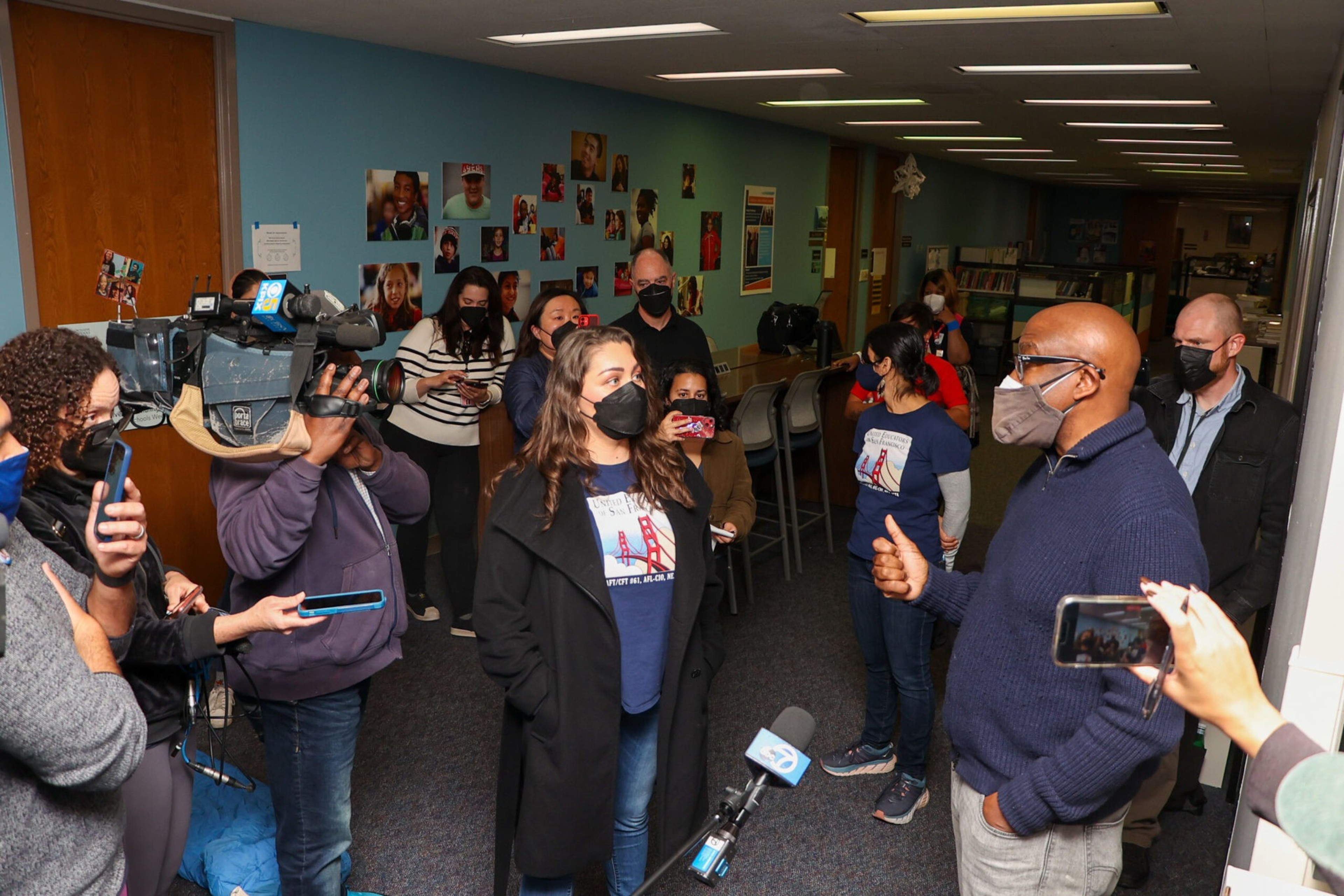 A group of masked people, some with cameras and microphones, interview a man giving a thumbs-up in a room with photos on the wall and stools at a counter.