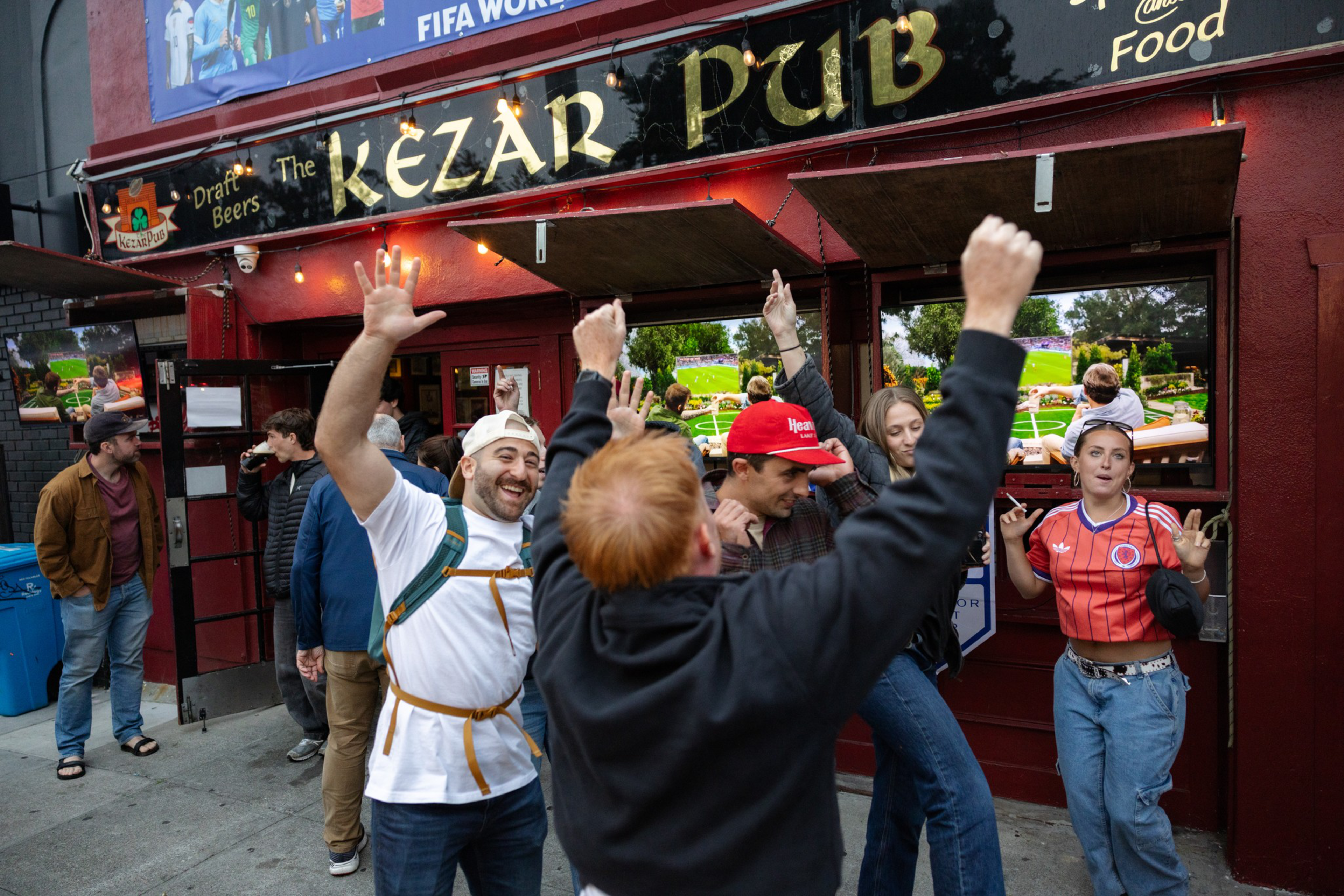 A group of people celebrates enthusiastically outside Kezar Pub, some raising their hands, with a sports game playing on multiple screens inside.
