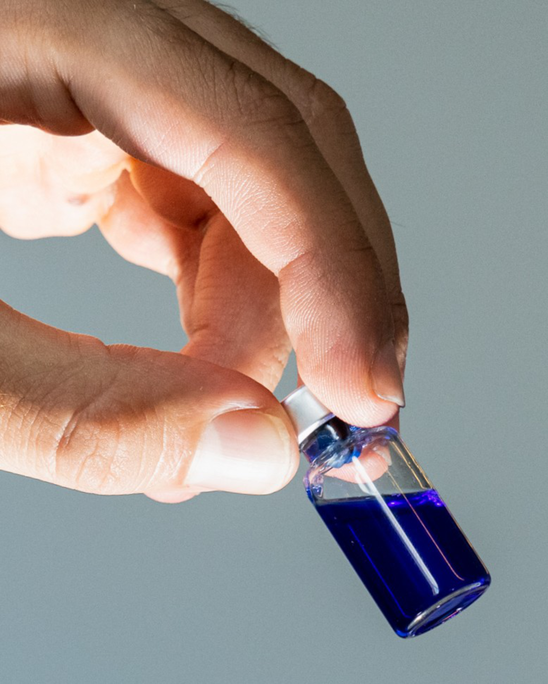 A hand gently holds a small glass vial filled about halfway with a dark blue liquid against a plain gray background.