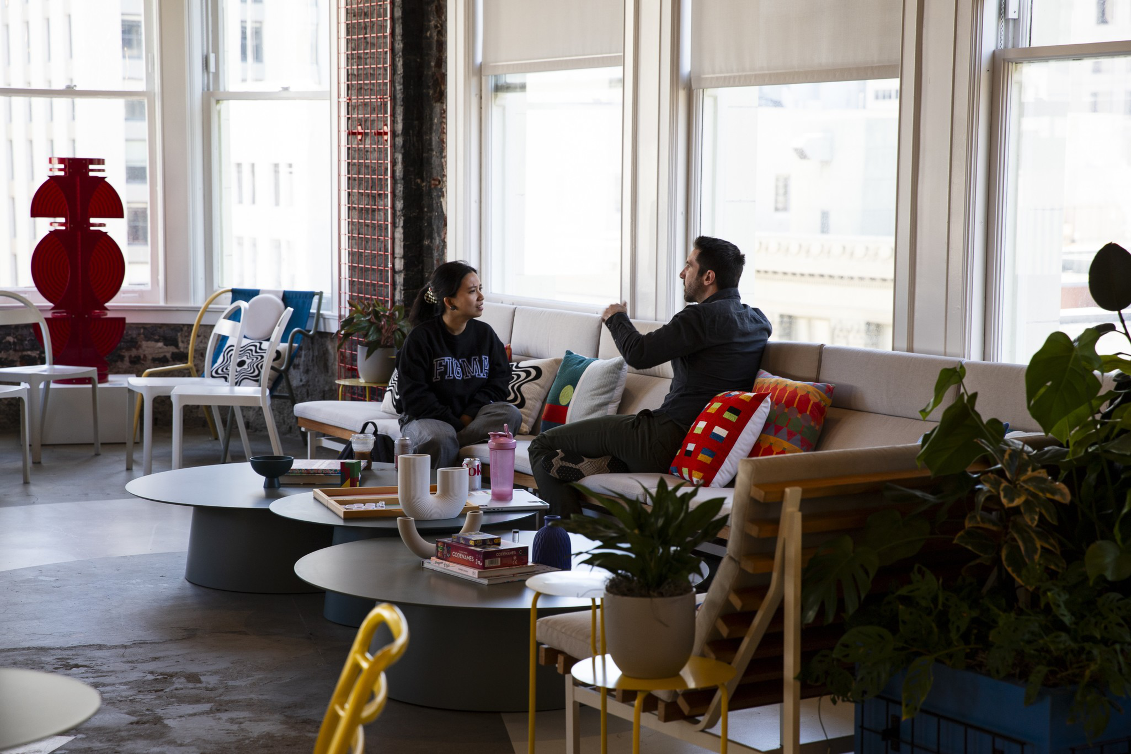 Two people sit and talk on a couch in a brightly lit room with large windows, colorful pillows, and modern decor including plants and abstract sculptures.