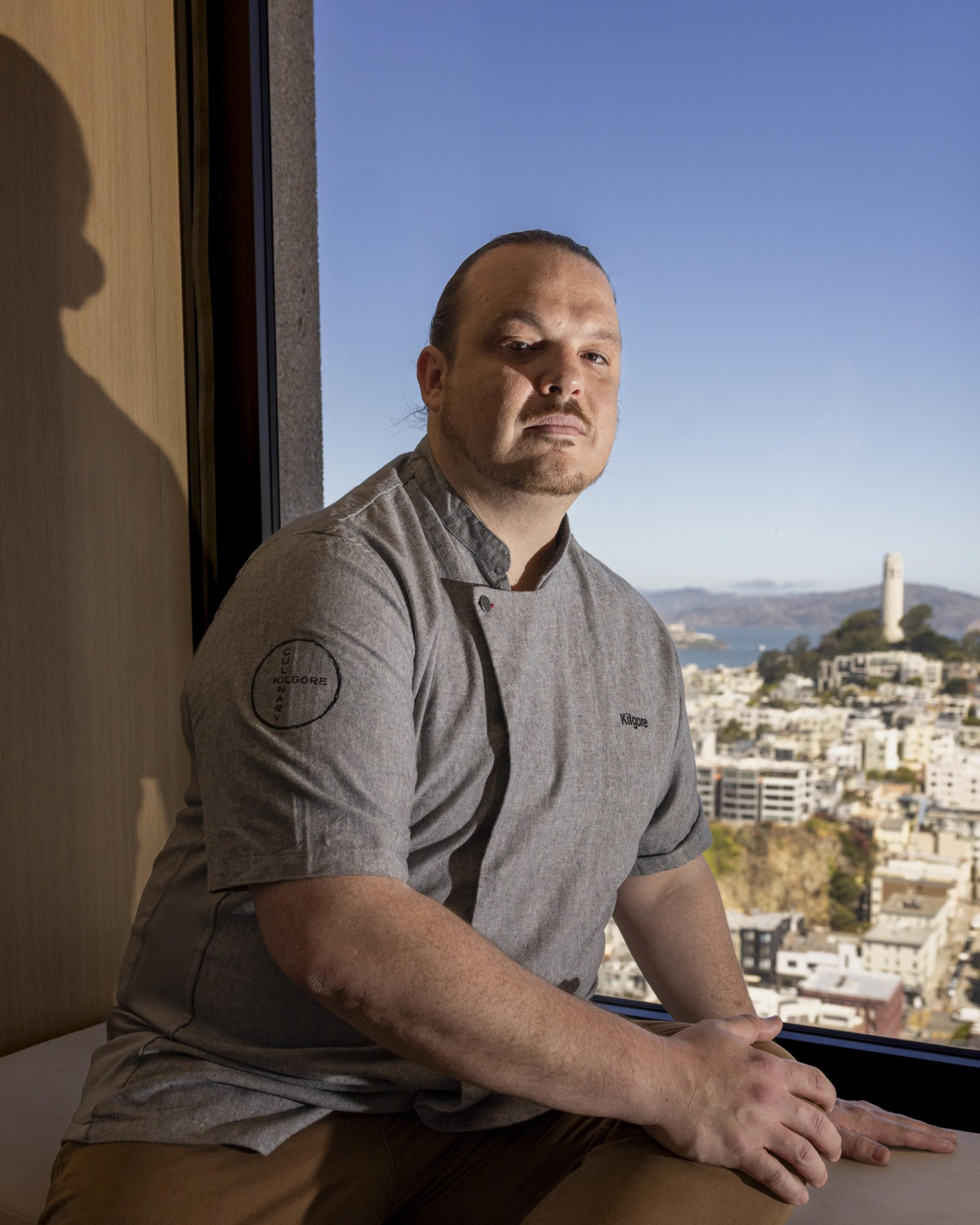 A man in a chef's coat sits by a large window with a cityscape and blue sky in the background. He is gazing intently at the camera with his hands resting on a surface.