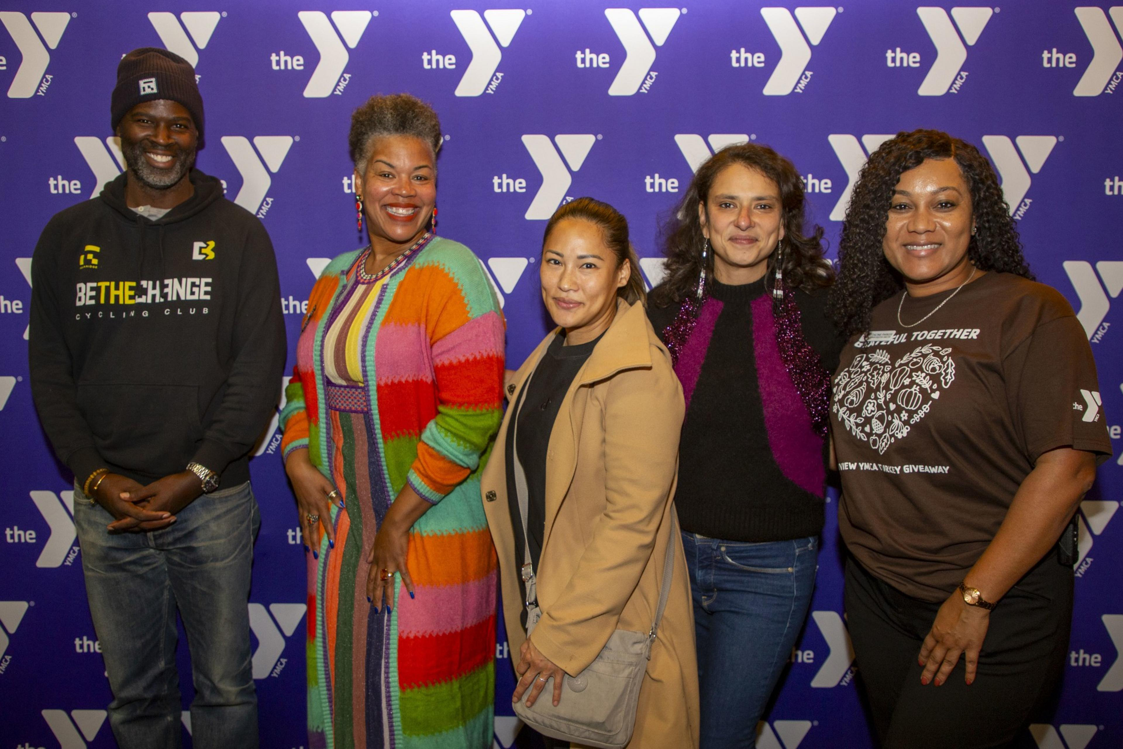 Five people stand smiling in front of a YMCA backdrop, with diverse styles ranging from a colorful coat to a brown t-shirt, capturing a sense of togetherness.