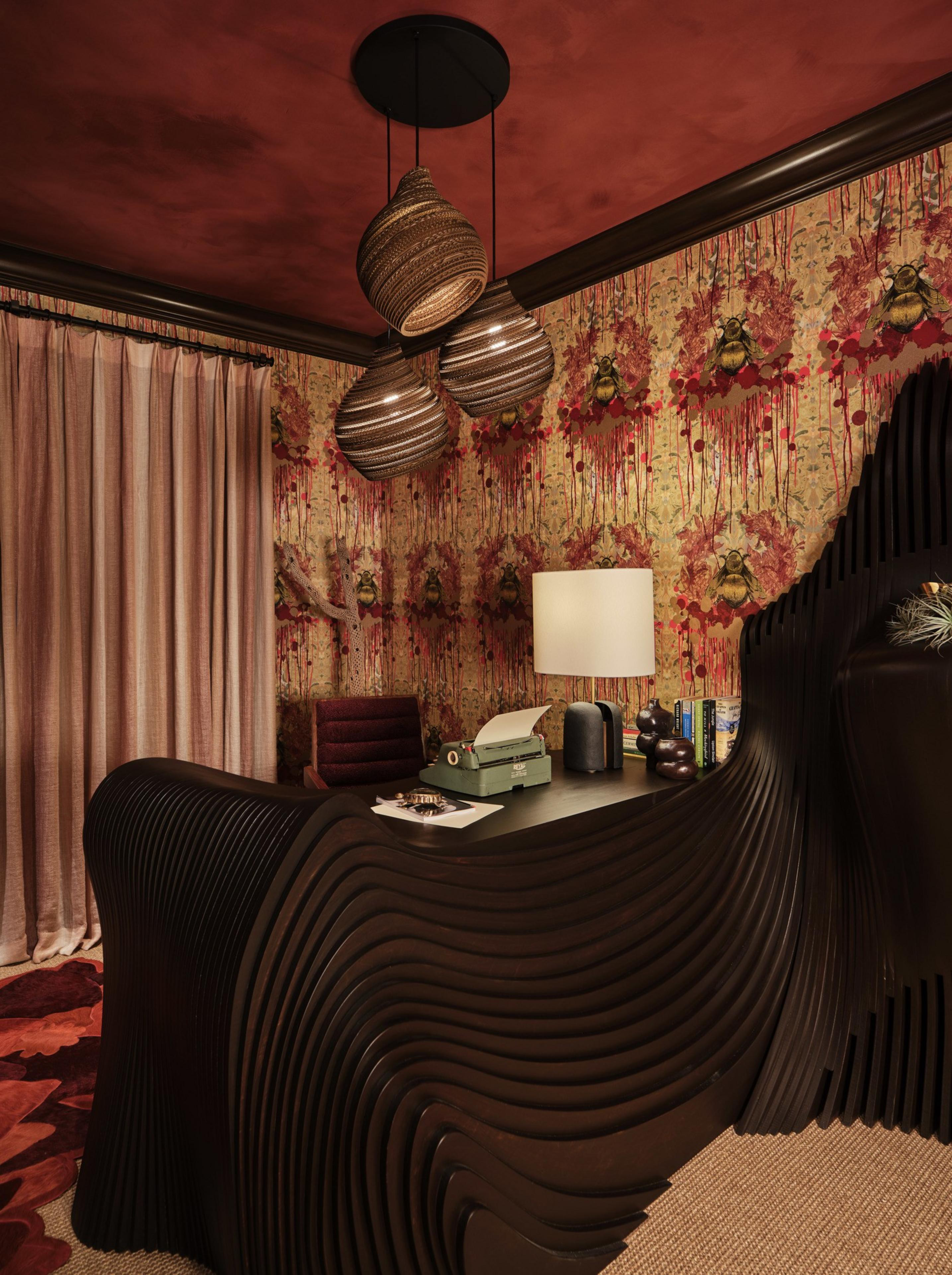 A dark wood desk with a wavy design sits in a room with red and yellow wallpaper featuring large bees, a typewriter, books, and a lamp on the desk.
