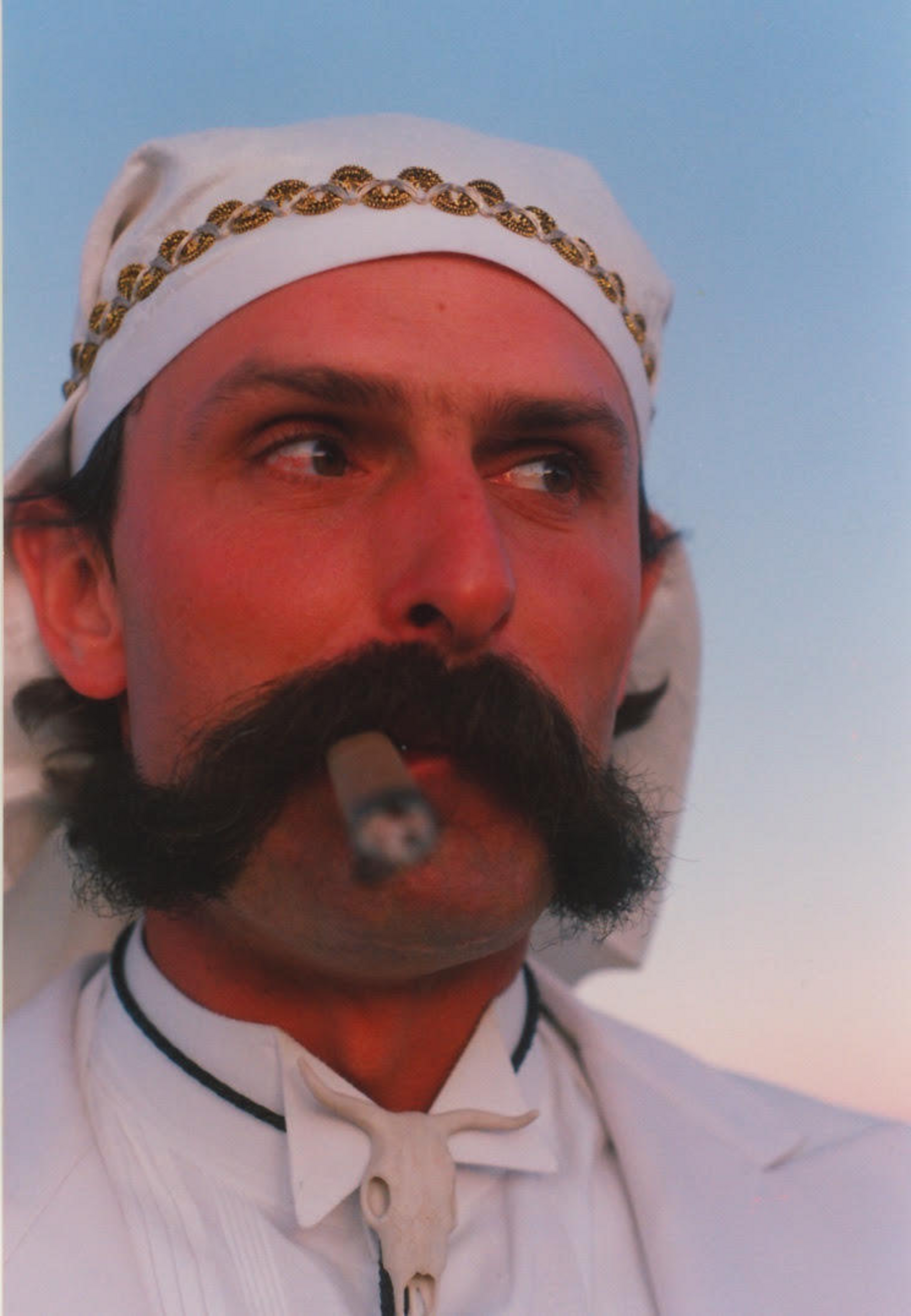 A man with a large dark mustache wears a white headscarf with gold trim, a white suit, and smokes a cigar while looking to the side.