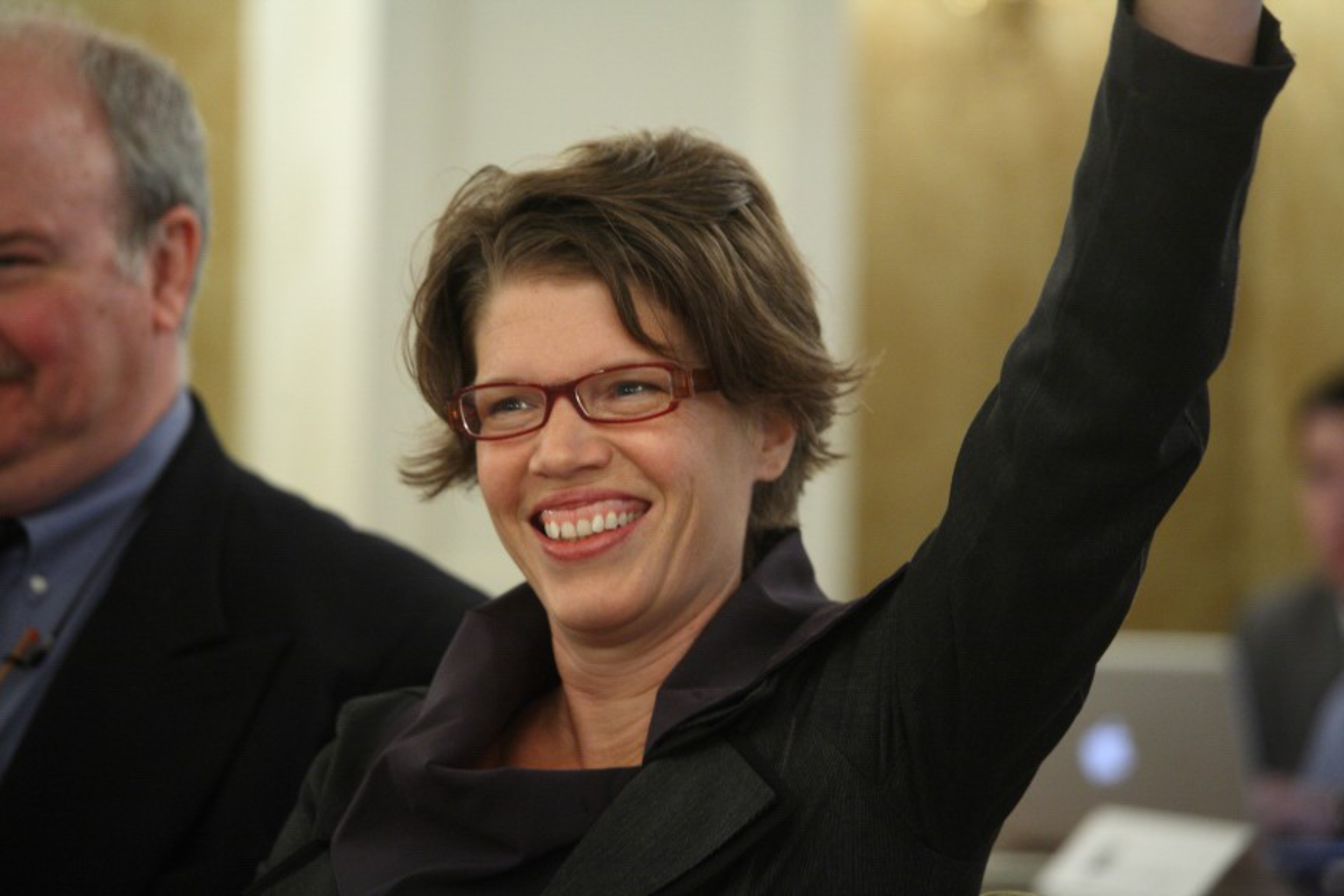 A smiling woman with short brown hair and red glasses raises her arm, wearing a dark jacket, with a blurred man and laptop in the background.