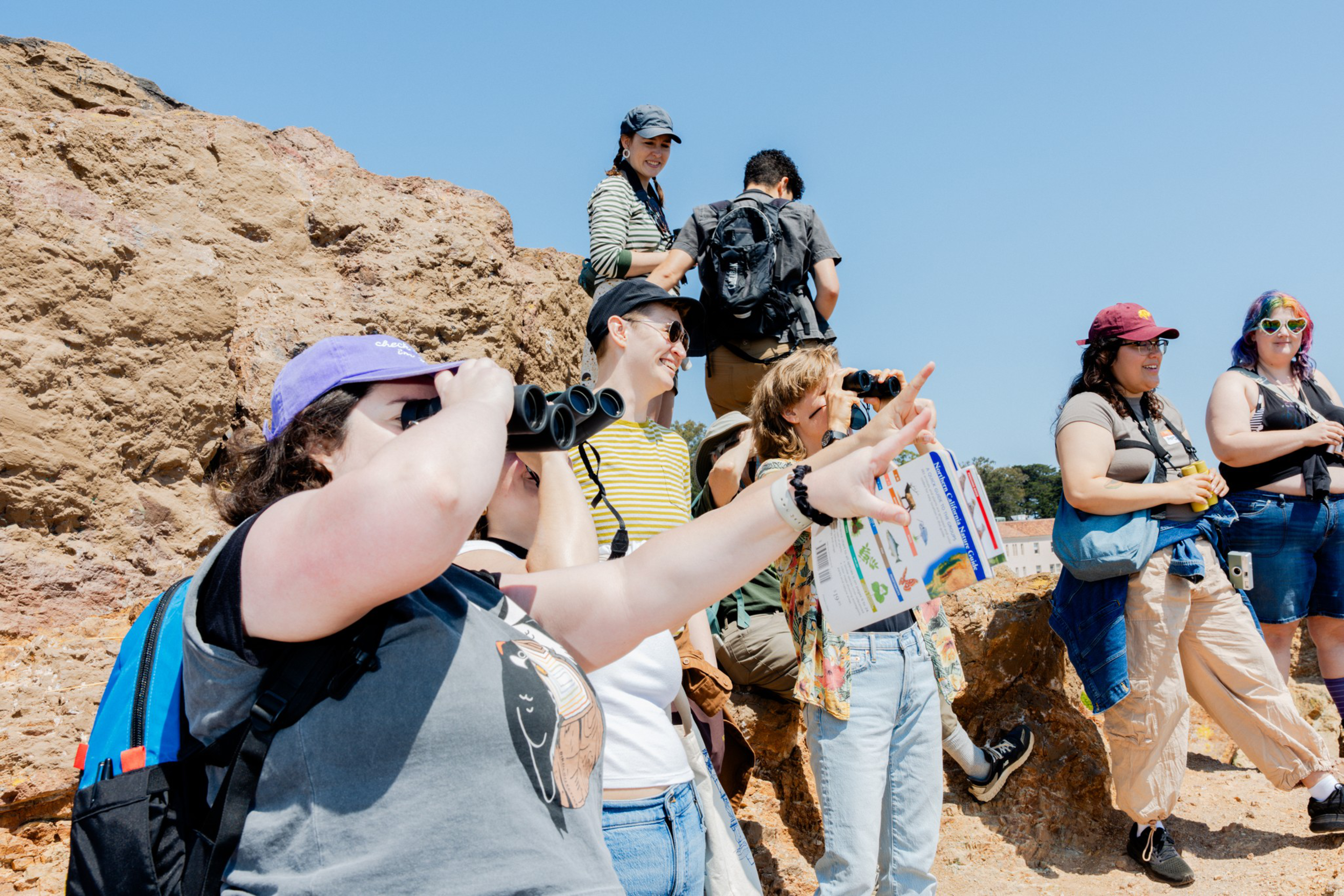 A group of people, some with binoculars and one pointing, are gathered on rocky terrain under a clear blue sky, engaging in outdoor observation.