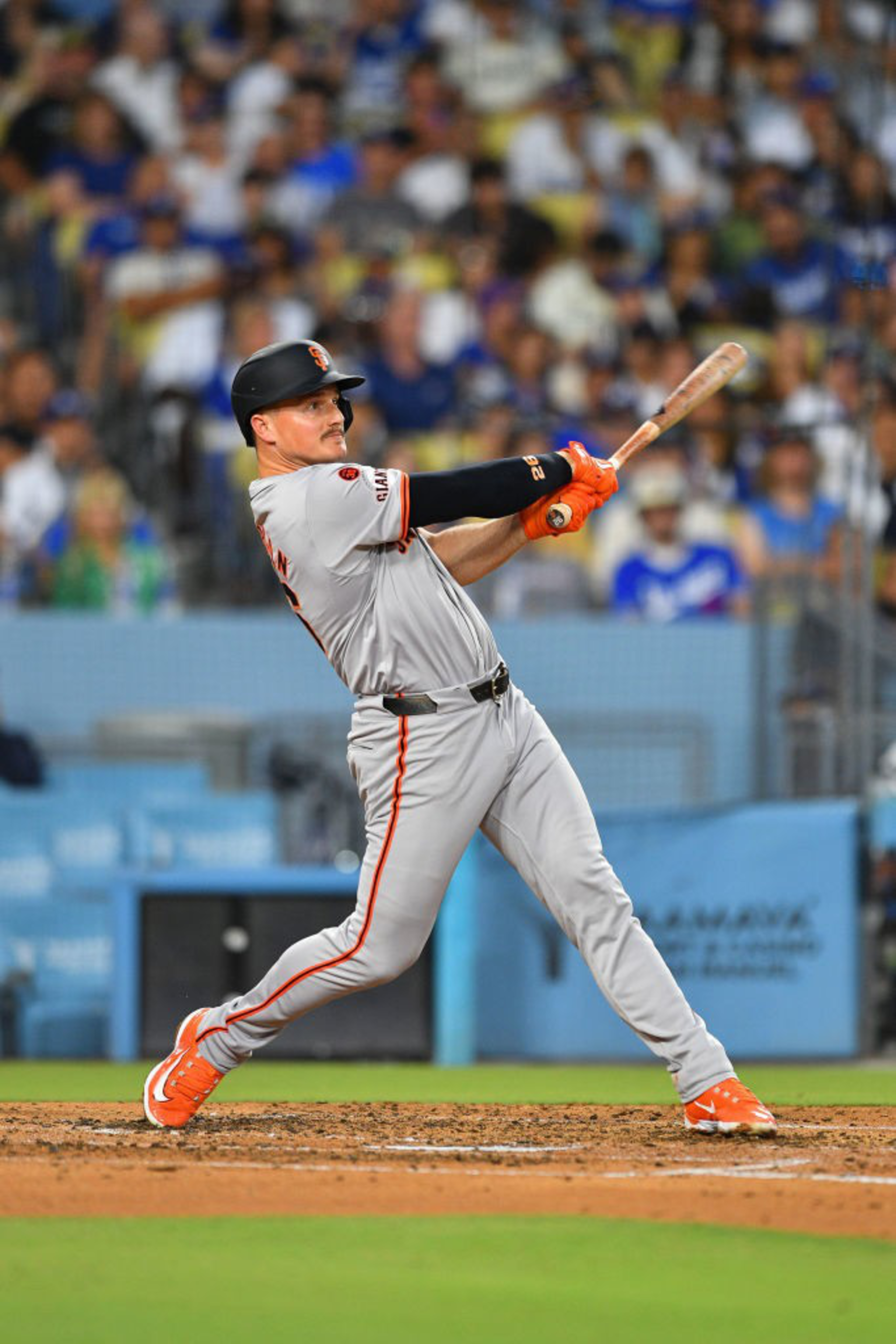 A baseball player in a gray and orange uniform is swinging a bat during a game. The background shows a crowd in a stadium with blurred spectators.
