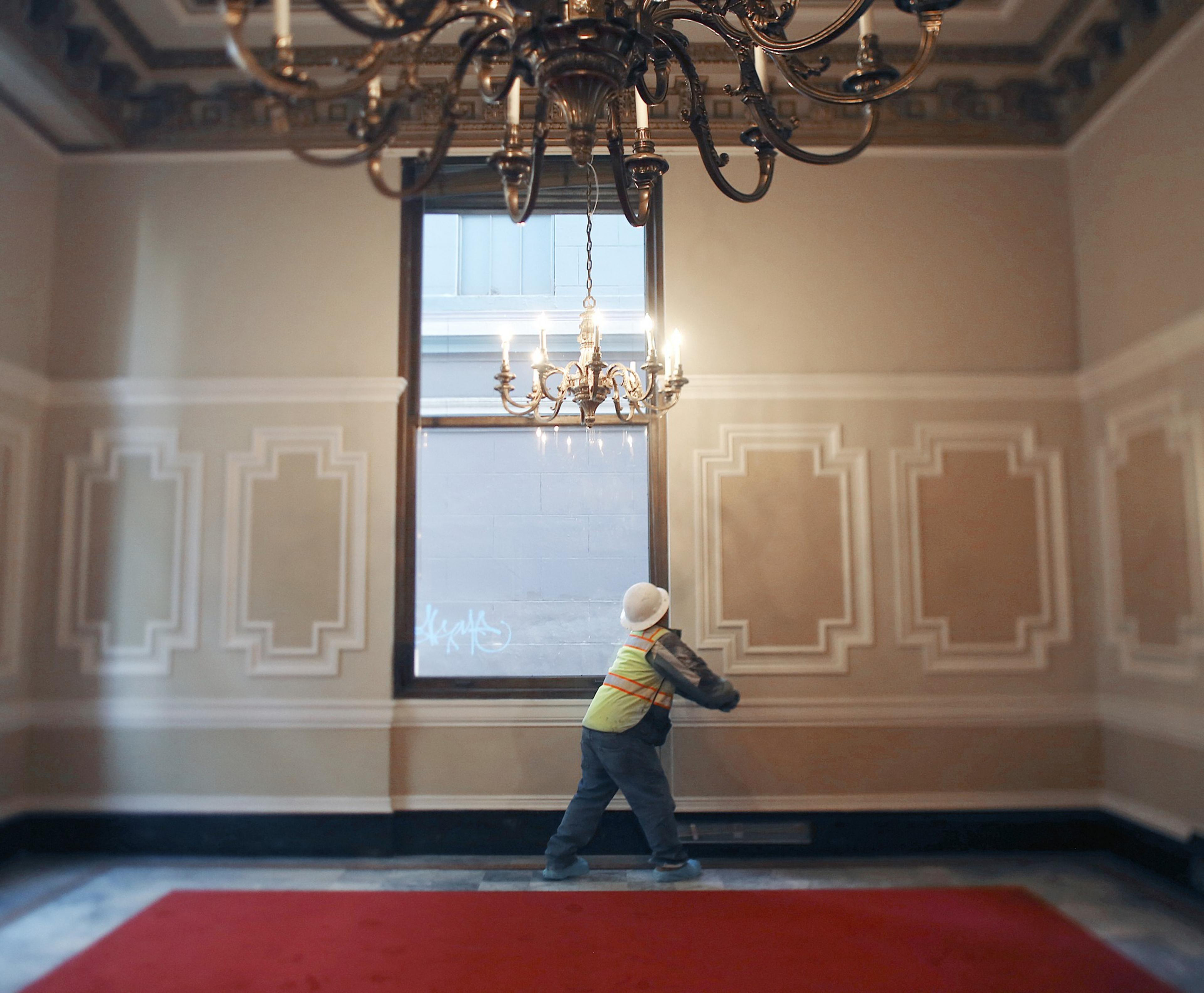 A worker in a hard hat and reflective vest is adjusting a window in an ornate room with detailed paneling and a large chandelier above.