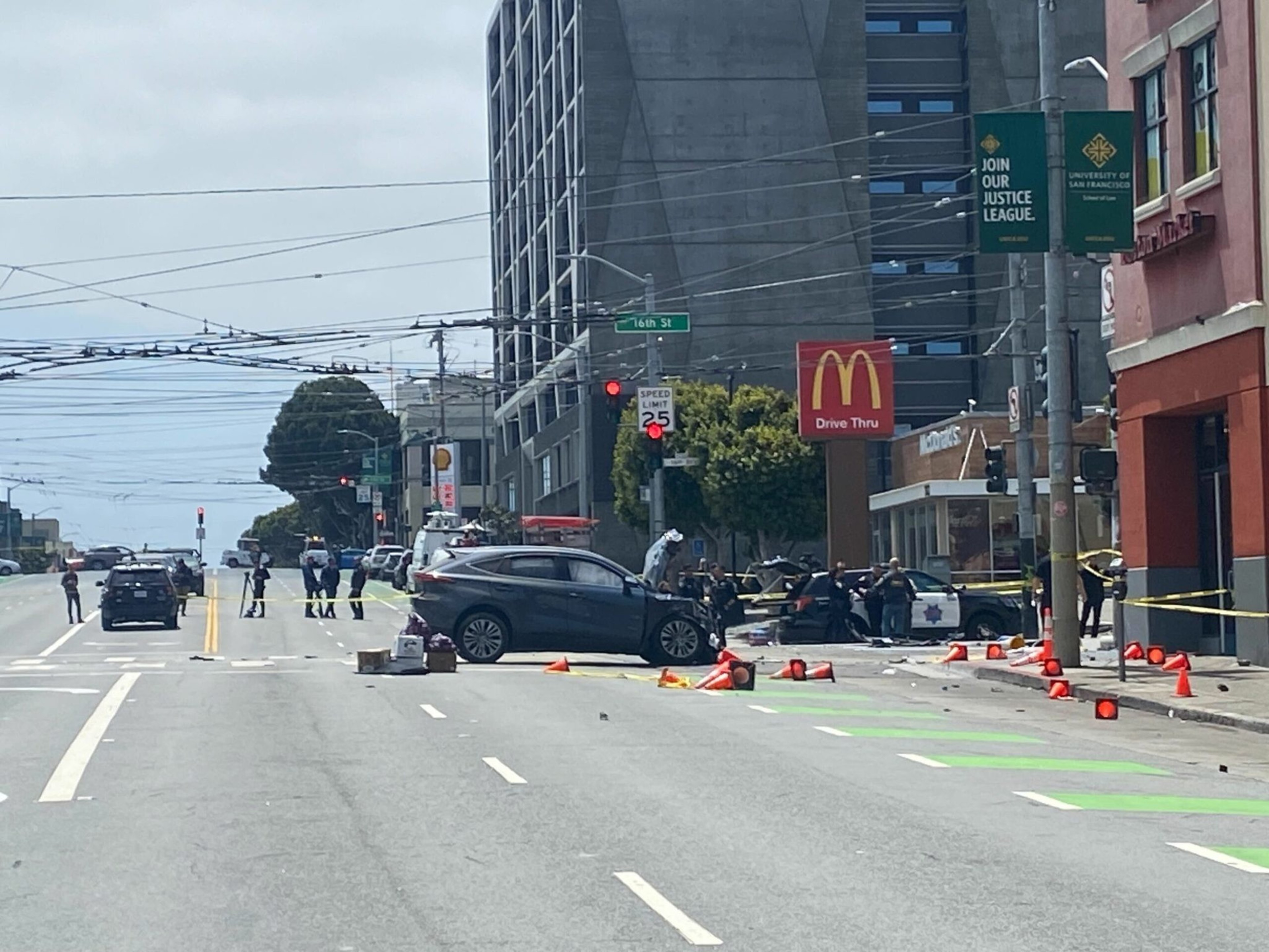 A dark SUV is stopped diagonally across the street with traffic cones scattered around and police officers gathered near a McDonald's drive-thru sign.