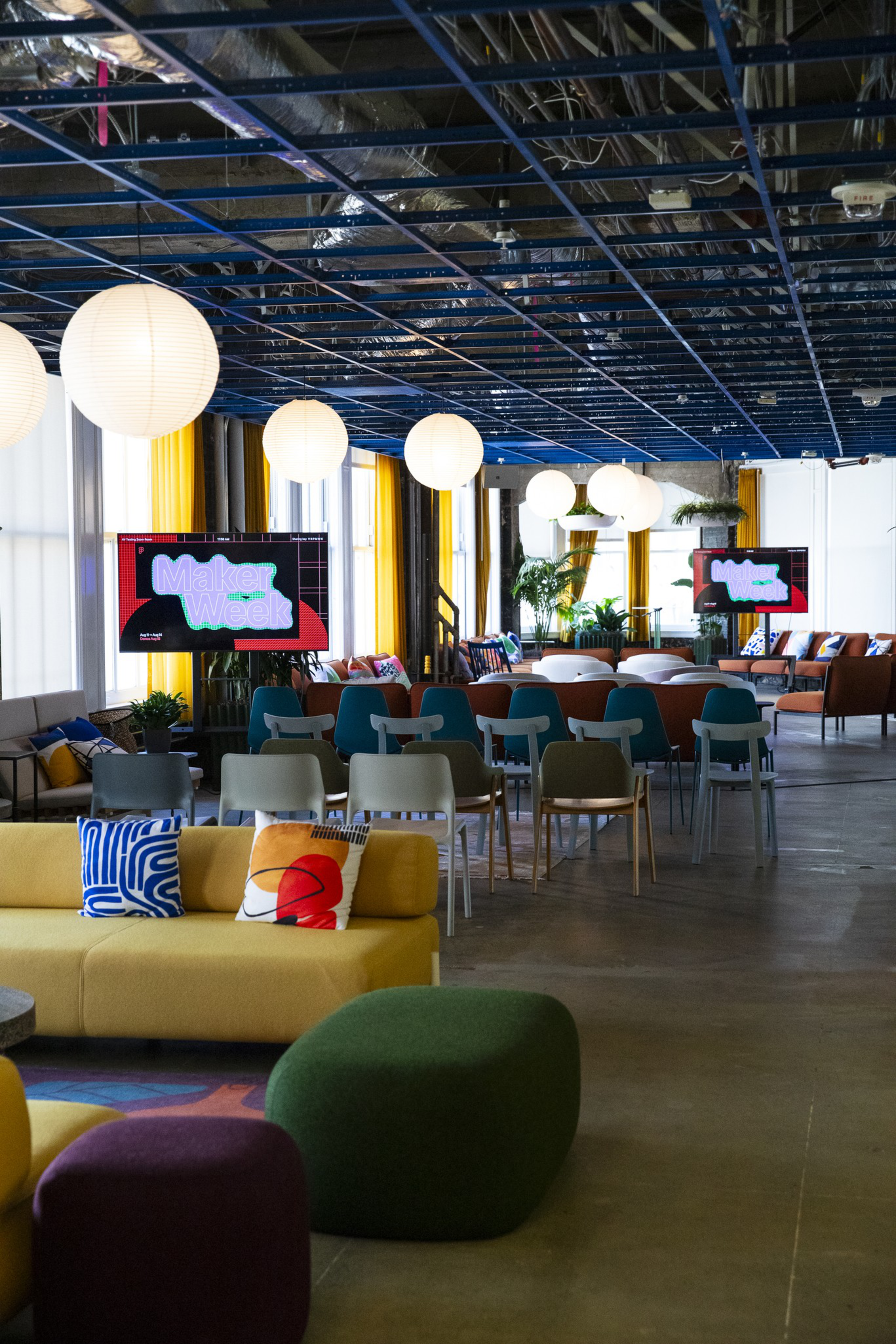 A brightly lit modern lounge space with colorful sofas, patterned pillows, multiple chairs, hanging round lights, and screens displaying "Maker Week."