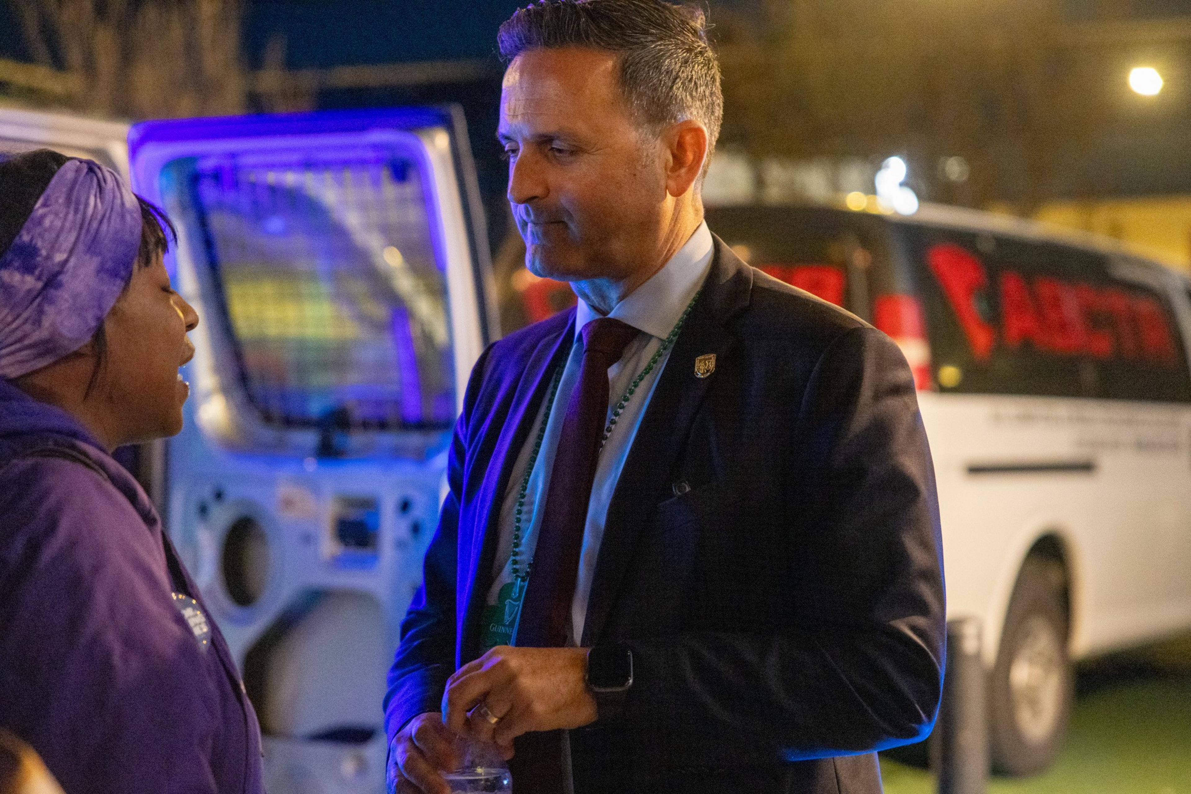 A man in a suit converses with a woman outside at night, near a lit-up ambulance.