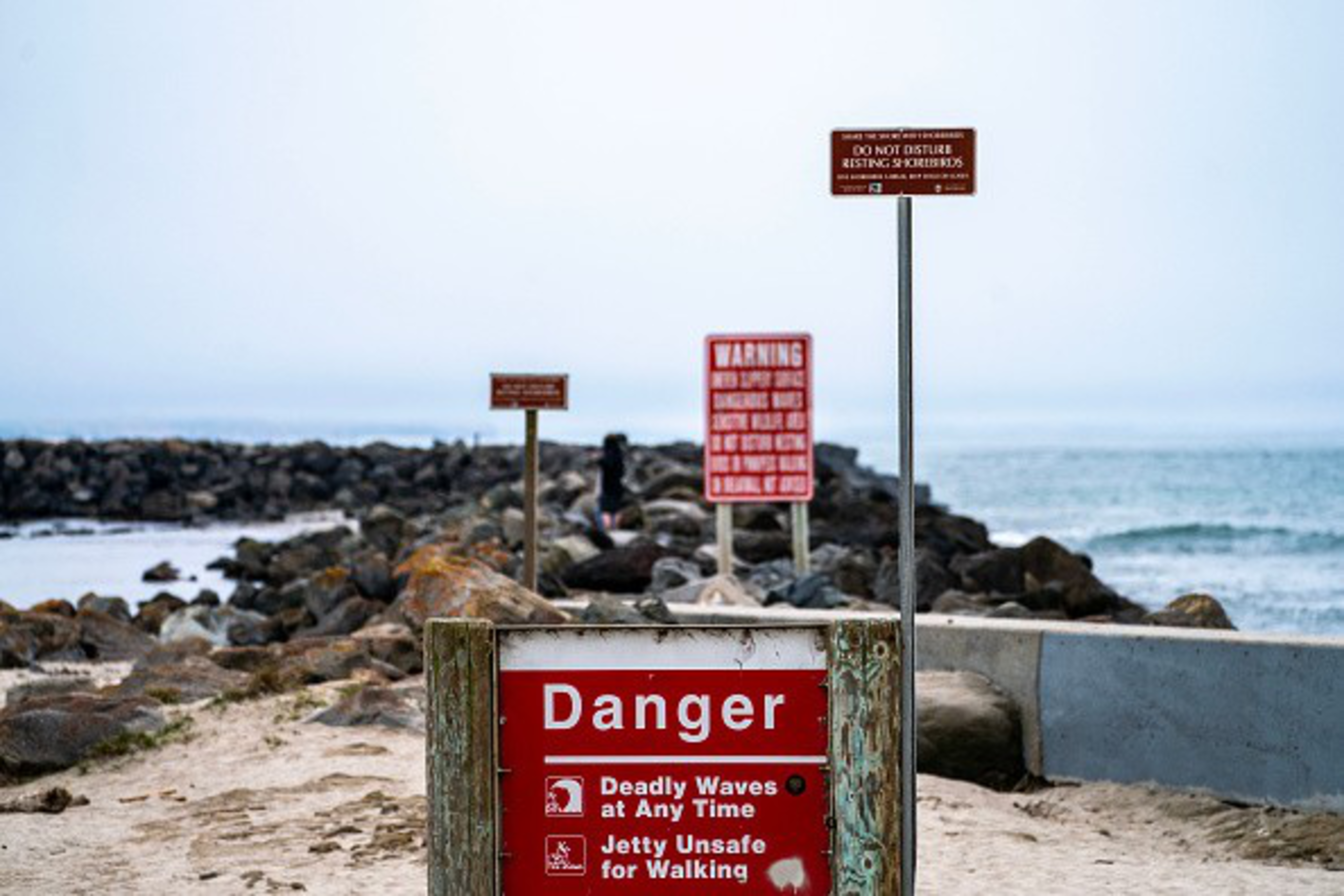A sandy path leads to a rocky jetty by the ocean, with multiple red warning signs stating danger and unsafe jetty conditions.