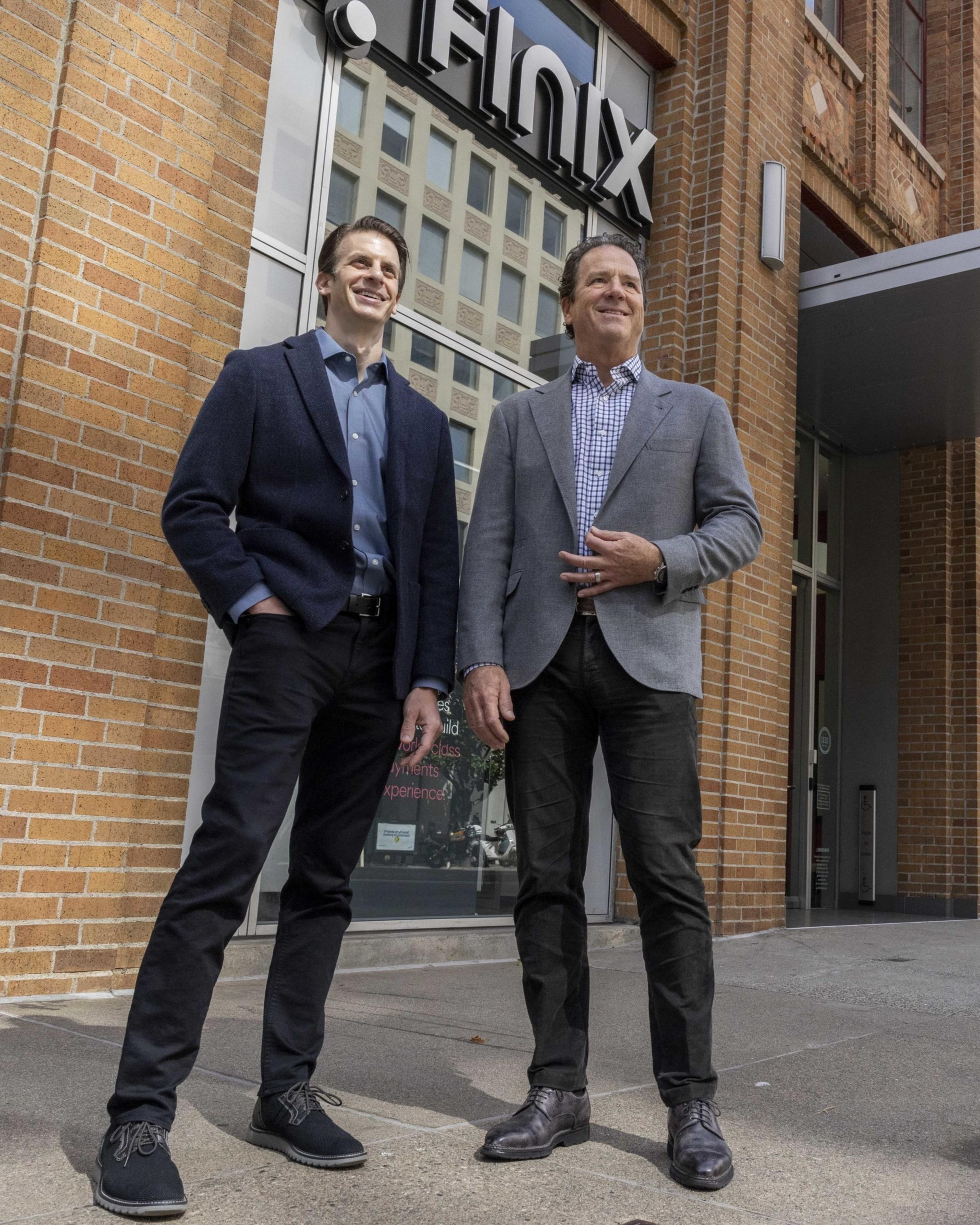 Two men, dressed in blazers and smiling, stand side by side in front of a building with a sign that reads "Finix."