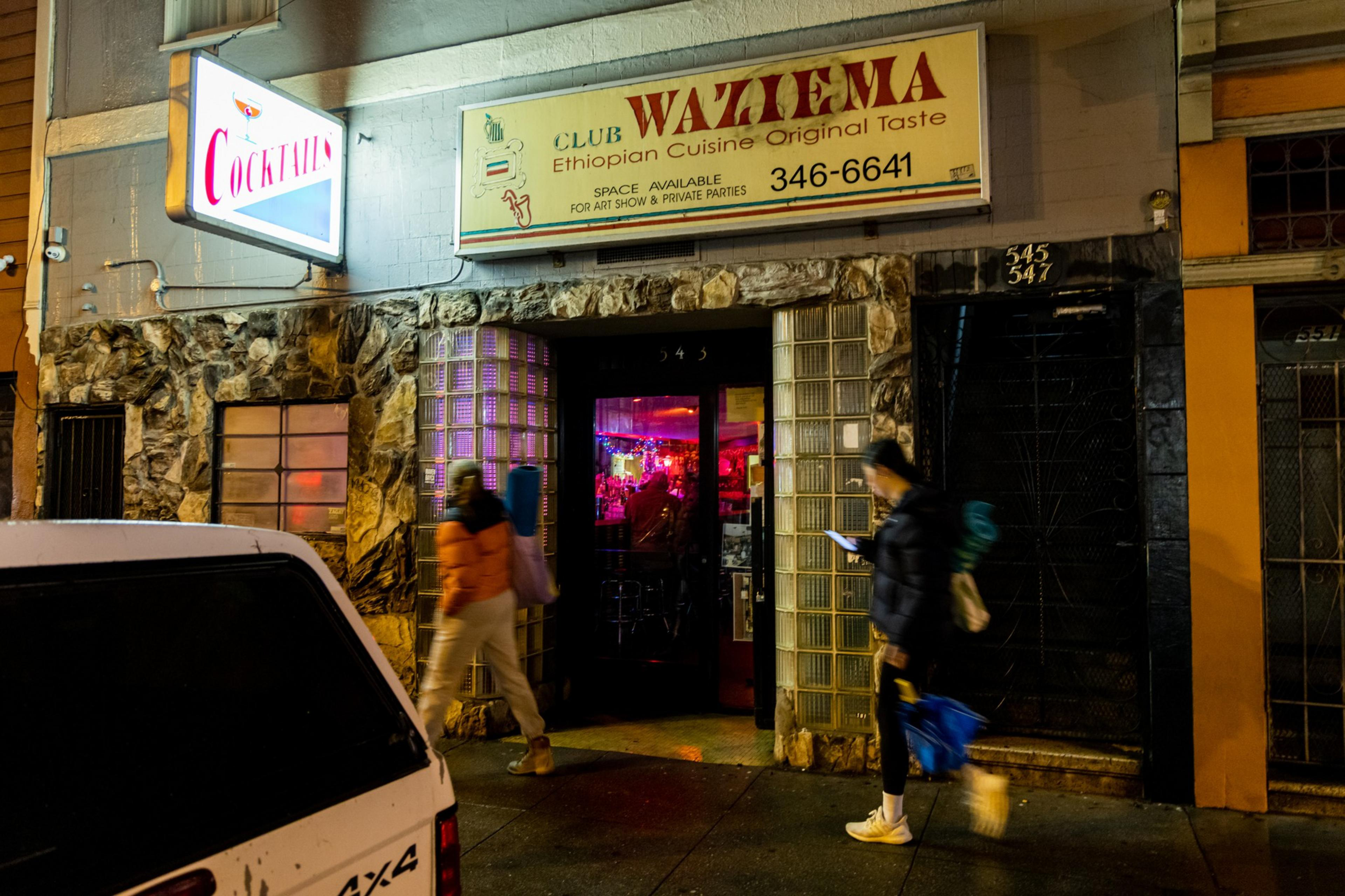 A dimly lit street scene shows a bar entrance with bright signs reading &quot;Cocktails&quot; and &quot;Waziema.&quot; Two people walk by, one glancing at a phone.